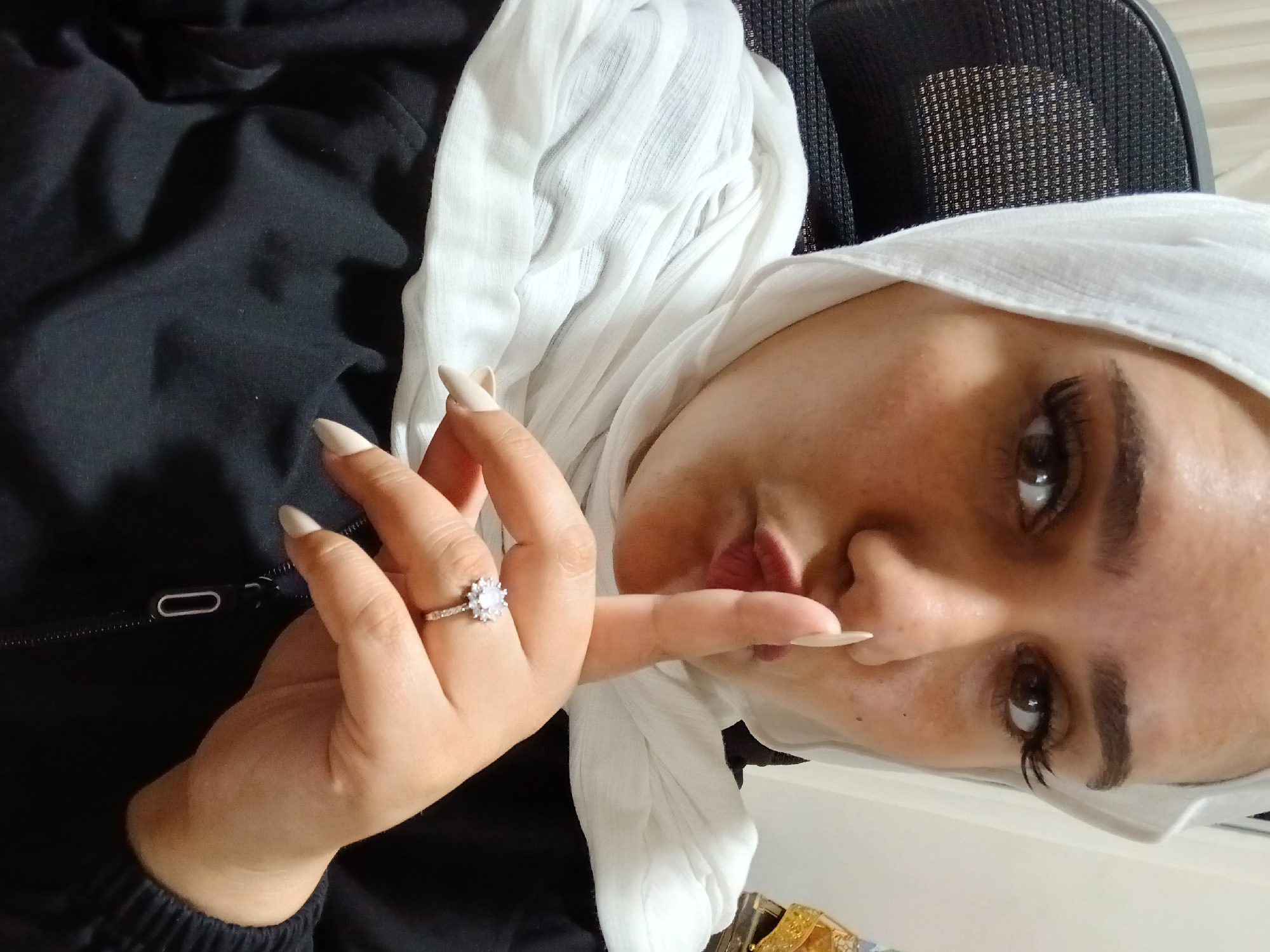 A woman wearing a white hijab and a black jacket is pictured from the chest up, looking directly at the camera with her lips pursed and one finger held to her lips in a shushing gesture. She has long, false eyelashes and is wearing a ring with a large, multifaceted stone surrounded by smaller stones on her right hand. The background appears to be an indoor setting with a textured chair back visible on the left and a blurred desk with some decorative items on the right. The overall lighting suggests an indoor environment, possibly an office or a home setting. The image does not provide any specific details that would allow for the localization of Rukn ad-Din Municipality, Syria.