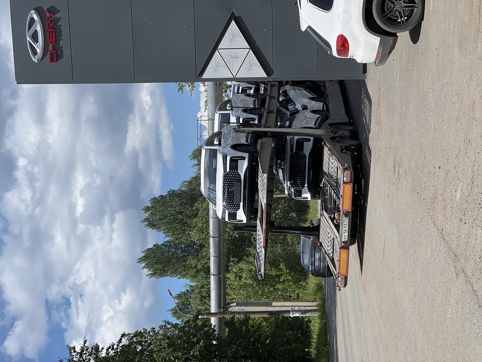 This image depicts a daytime scene at a Chery car dealership, likely in Izhevsk, Russia, evidenced by the "18" region code on a parked vehicle's license plate. A multi-level car carrier is positioned next to the dealership building, seemingly in the process of delivering new vehicles.
The prominent Chery dealership building, located on the right side of the frame, features a grey facade with a large, polished chrome Chery logo and the brand name "CHERY" rendered in red letters below it. To the right and below the logo, a decorative white arrow-shaped art installation composed of stacked triangular segments is affixed to the building.
The car carrier, a large dark grey articulated truck trailer, is loaded with at least four new white SUVs. The top level clearly displays two Chery Tiggo 8 Pro Max SUVs, identifiable by their distinctive front grilles, headlights, and daytime running lights. The bottom level holds at least two more similar white SUVs, partially obscured. The rear of the car carrier displays yellow and red reflective markers and a Russian license plate "YY 1613 77".
In the mid-ground, behind the car carrier, a dark grey Hyundai sedan (model appears to be a Solaris or Accent) is parked on an asphalt road, partially obscured by the trailer. Its rear license plate, "E 628 HT 18", confirms the vehicle is registered in the Udmurt Republic, where Izhevsk is the capital.
The background on the left features a dense line of tall green deciduous trees under a bright blue sky with scattered white cumulus clouds. A utility pole with a streetlamp and overhead wires stands to the left of the car carrier. A large, horizontal grey pipeline, supported by poles, traverses above the road. The ground consists of asphalt leading to a grassy verge on the left. No individuals are visible in the frame.