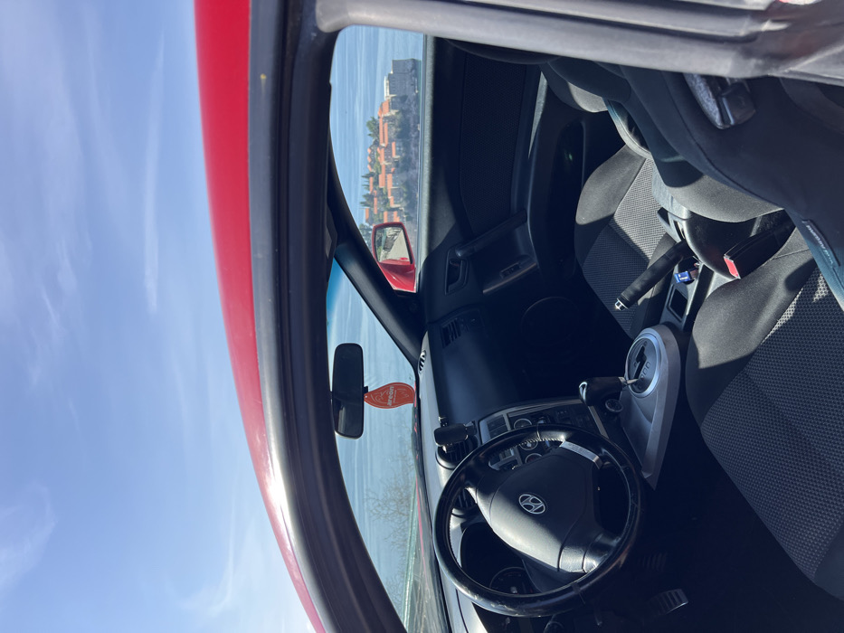 The image depicts the interior of a red car, specifically from the perspective of an observer standing outside the open driver's side door, looking inward and outward. The car's interior, primarily dark grey and black, includes a multi-function steering wheel with a Hyundai logo, a dashboard, and two front bucket seats upholstered in dark grey fabric. An automatic gear shifter is located on the center console, alongside a raised handbrake lever. A phone mount is affixed to the dashboard, and an orange "Areon" brand air freshener hangs from the rearview mirror.
Through the car's windshield and passenger side window, the distinctive fortified village of Sveti Stefan, Montenegro, is clearly visible situated on a rocky islet in a calm, blue body of water, likely the Adriatic Sea. The village features numerous red-tiled roofs and stone buildings. The sky above is clear and blue with minimal cloud cover. The overall scene suggests a bright, sunny day. No individuals are present within the frame.