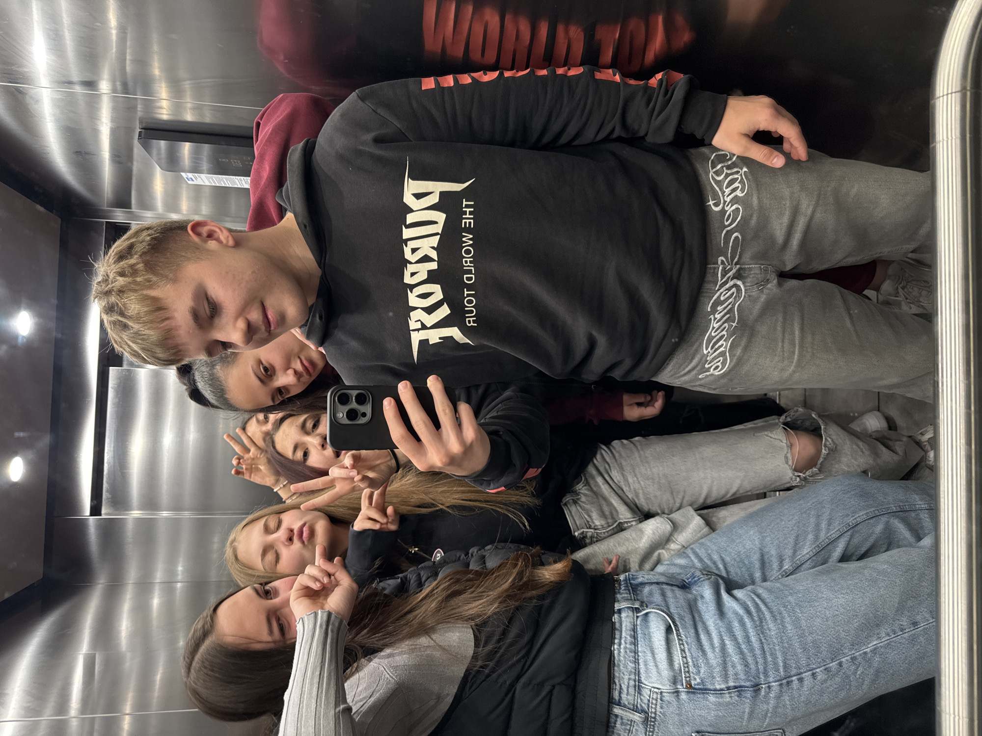 The image captures a group of five young individuals taking a mirror selfie inside an elevator in Sliven, Bulgaria. The setting is an enclosed space with polished, reflective metallic walls that show overhead lighting and the group's reflections.
On the left side of the frame, a young man with short blonde hair is visible, wearing a black hoodie. The hoodie features the reversed text "PURPOSE" in a stylized font and "THE WORLD TOUR" underneath. He is also wearing light grey jeans with a prominent white, script-like design running vertically down the outer seam. Directly behind him, another person is partially visible.
Towards the center and right, several young women are present. One holds a smartphone (an iPhone model with multiple cameras) to capture the selfie. Other women are making peace signs with their fingers, and one is seen pouting her lips playfully. Their attire includes a grey ribbed long-sleeve top, a black puffer vest, and various denim jeans, including light blue and ripped styles.
The activity is a casual group photo session, with the subjects exhibiting relaxed and friendly expressions. Faint reflections of the "WORLD TOUR" text from the hoodie are also visible on the upper part of the metallic wall. A small, dark panel with an unreadable digital display is mounted on the elevator wall.