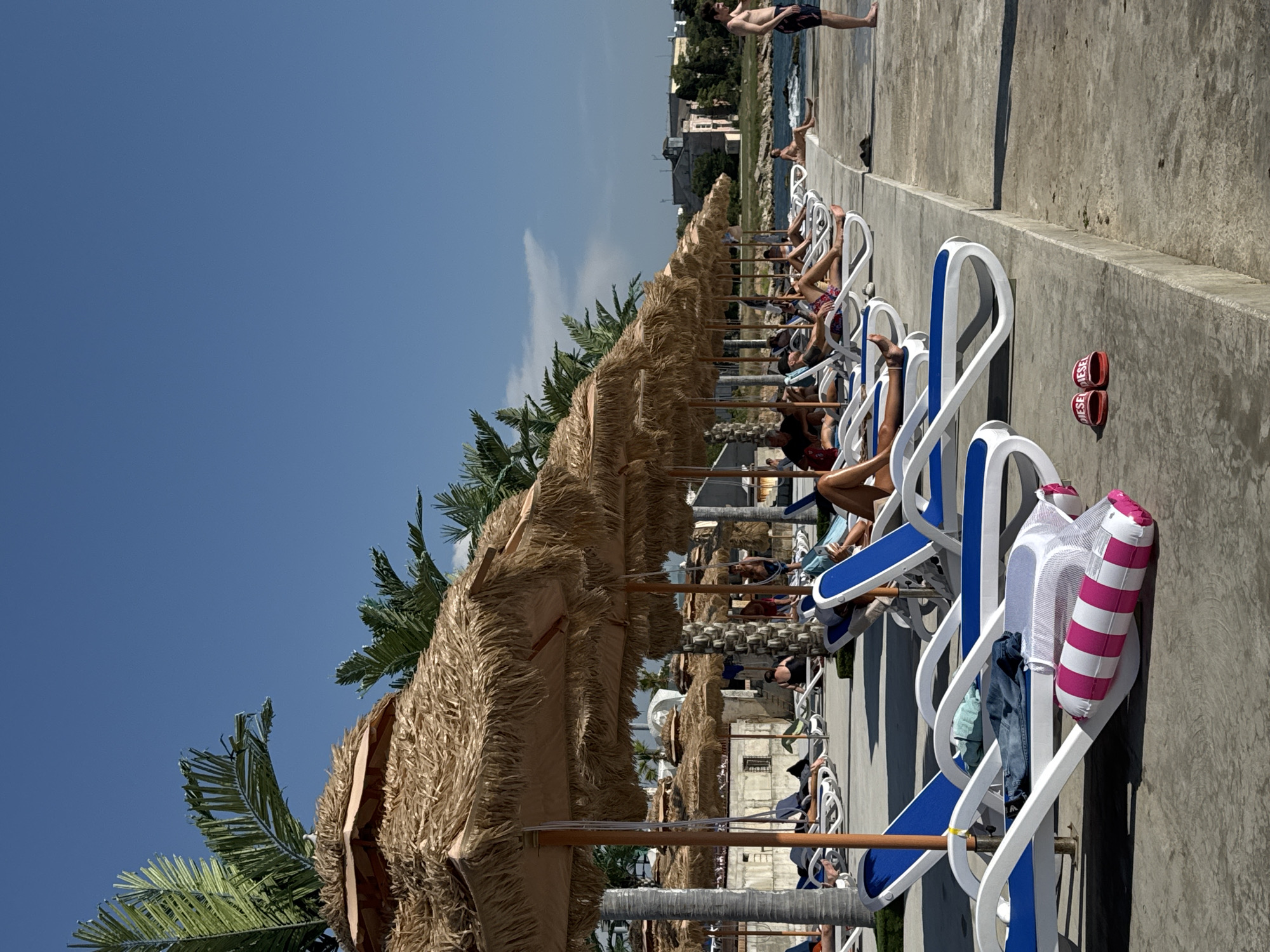 A row of thatched umbrellas and lounge chairs are set up on a concrete promenade along the Black Sea in Sevastopol, Ukraine. Several people are relaxing on the chairs, some reading and others sunbathing. Palm trees are visible behind the umbrellas, and a building with a white dome is in the background. The sky is clear and blue, and the sun is shining. In the foreground, a pair of red flip-flops rest on the concrete, and a striped inflatable toy lies on a lounge chair.