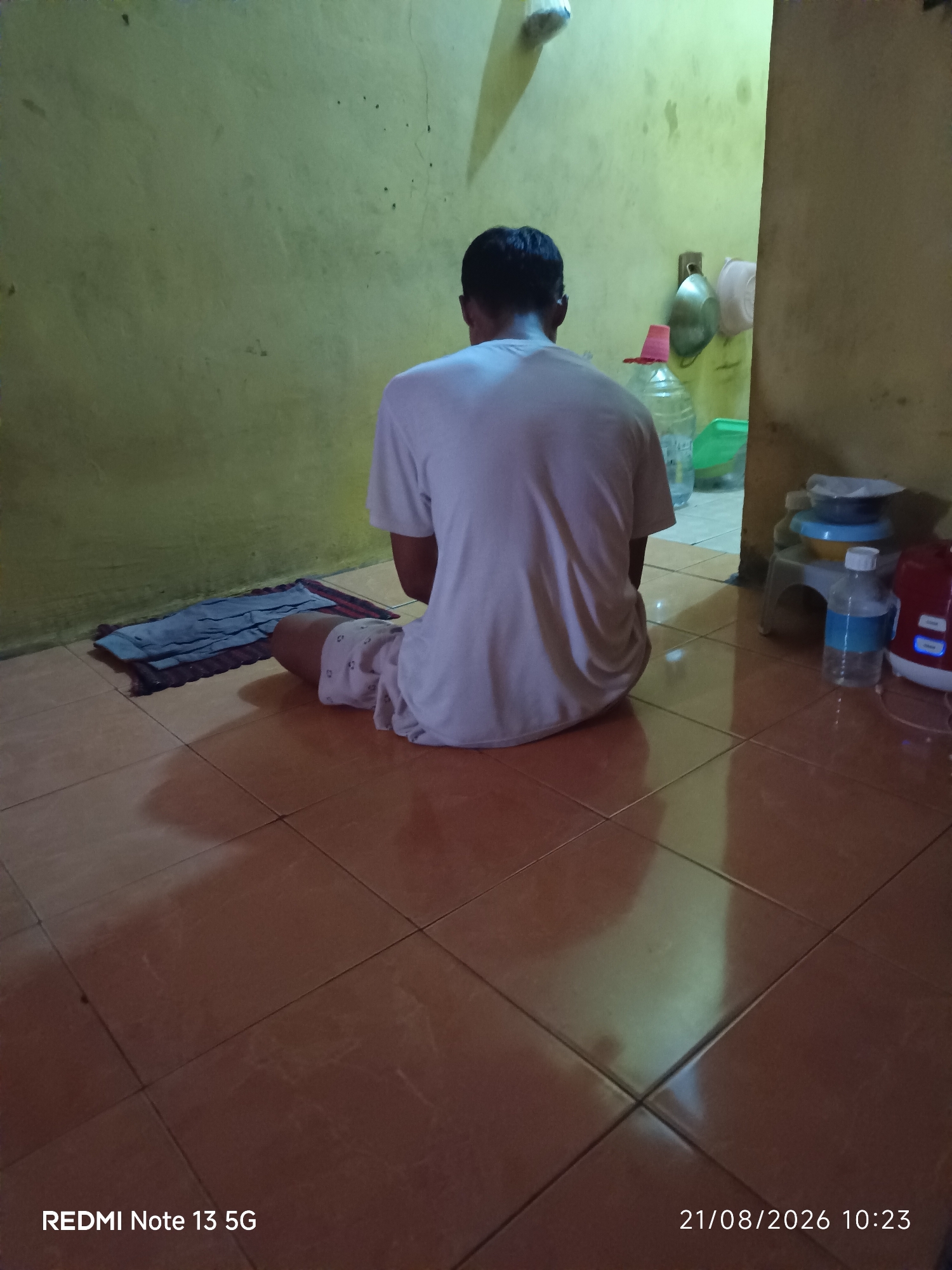 A person is seen from behind, kneeling on a prayer mat on a tiled floor. They are wearing a white t-shirt and light-colored pants with a pattern. The room has yellow walls and a brown tiled floor that reflects the light. To the left of the person is a blue prayer mat. To the right, there are various objects, including plastic bottles, containers, and a red appliance that appears to be a rice cooker. The lighting in the room is dim and casts shadows. The timestamp in the bottom right corner indicates "21/08/2026 10:23", and the text "REDMI Note 13 5G" is present in the bottom left corner. This scene is in Talaga Sari, Indonesia.