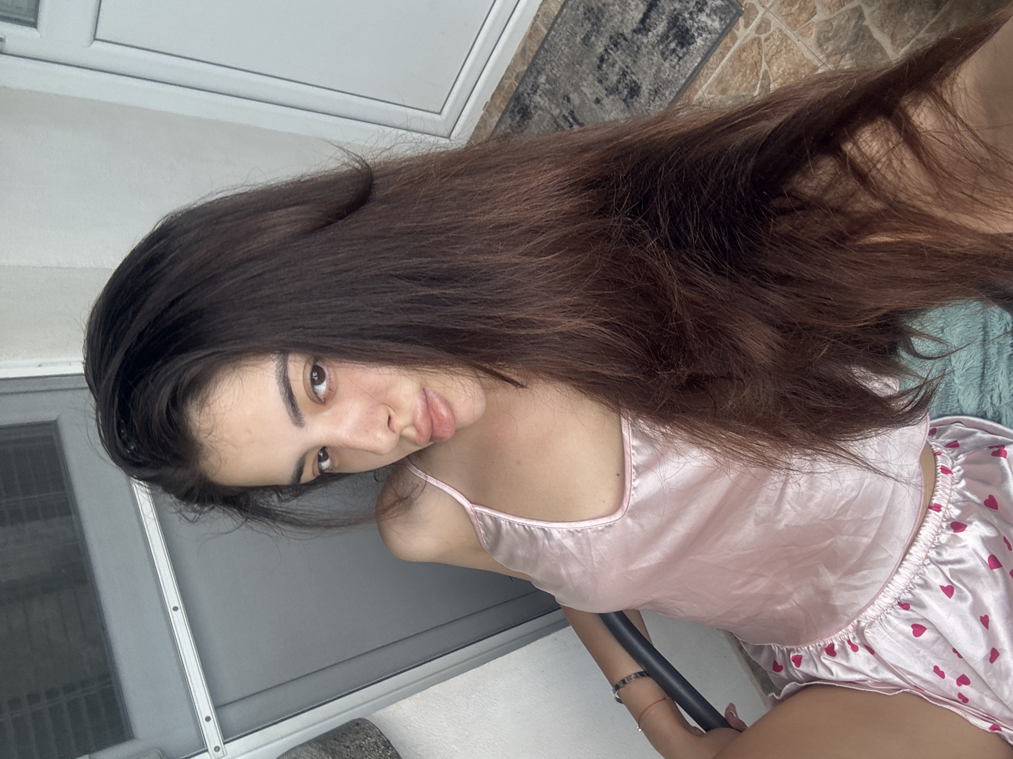 A young adult female is depicted in a medium shot, seated indoors or on a balcony. She has long, dark brown hair with reddish undertones, which appears slightly disheveled and falls over her left shoulder and extends across her torso. She wears a light pink, satin-like camisole with thin spaghetti straps and matching light pink shorts adorned with red heart patterns. Her right arm is visible, resting on a dark-colored, possibly metallic, armrest or railing. A silver-toned bracelet and a red string bracelet are on her right wrist. Her head is tilted slightly to her right, and her gaze is directed towards the camera. Her lips are slightly parted in a pout.
The background on the left features a white, textured wall adjacent to a grey window or door frame with a screen or horizontal blinds visible behind it. On the right, a white PVC door or window frame is visible against a light wall. The floor beneath the right frame consists of irregularly shaped, light brown and beige tiles, possibly indicating an outdoor or balcony setting. A grey patterned rug or mat lies partially on these tiles. A light blue or teal fuzzy textile, possibly a blanket or rug, is positioned beneath the subject's left leg. The lighting suggests natural daylight. The scene is situated in Plovdiv, Bulgaria.