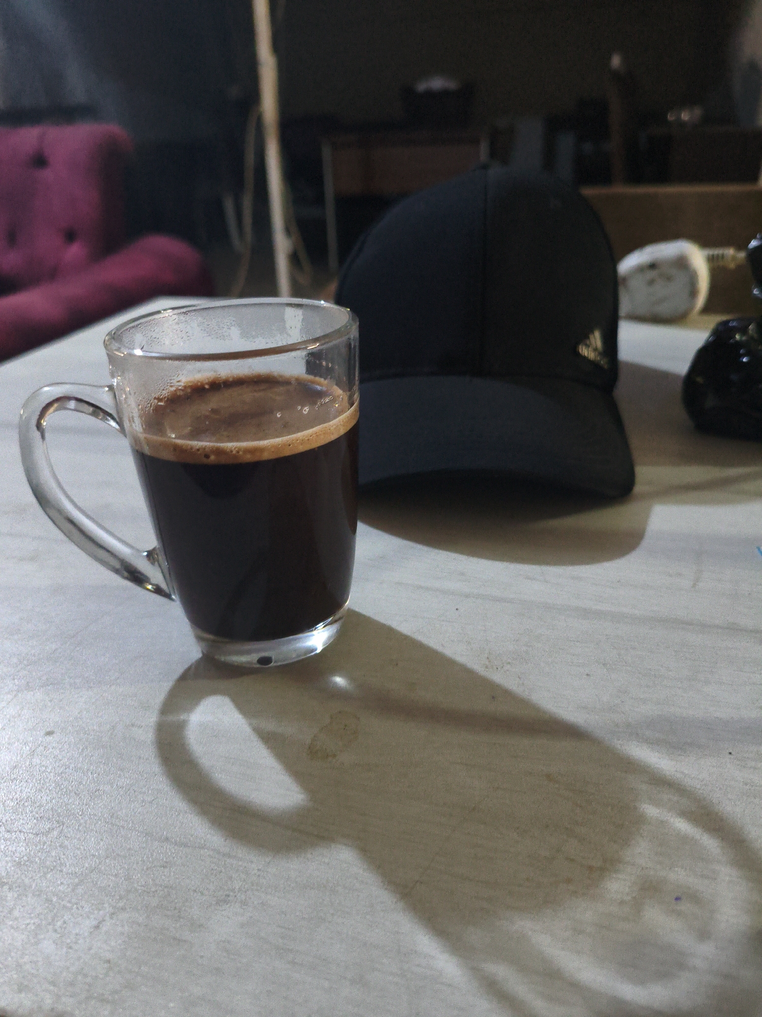An Adidas baseball cap is placed on a table next to a glass mug of dark liquid. The mug is partially filled and has a foam layer on top. The cap is black and has the Adidas logo on the front. The table appears to be made of wood and is lightly textured. The background is dark and out of focus, but a pink sofa and some vertical elements are visible. The scene is lit by a light source that casts shadows of the mug and cap onto the table. The image appears to be taken in an indoor setting.