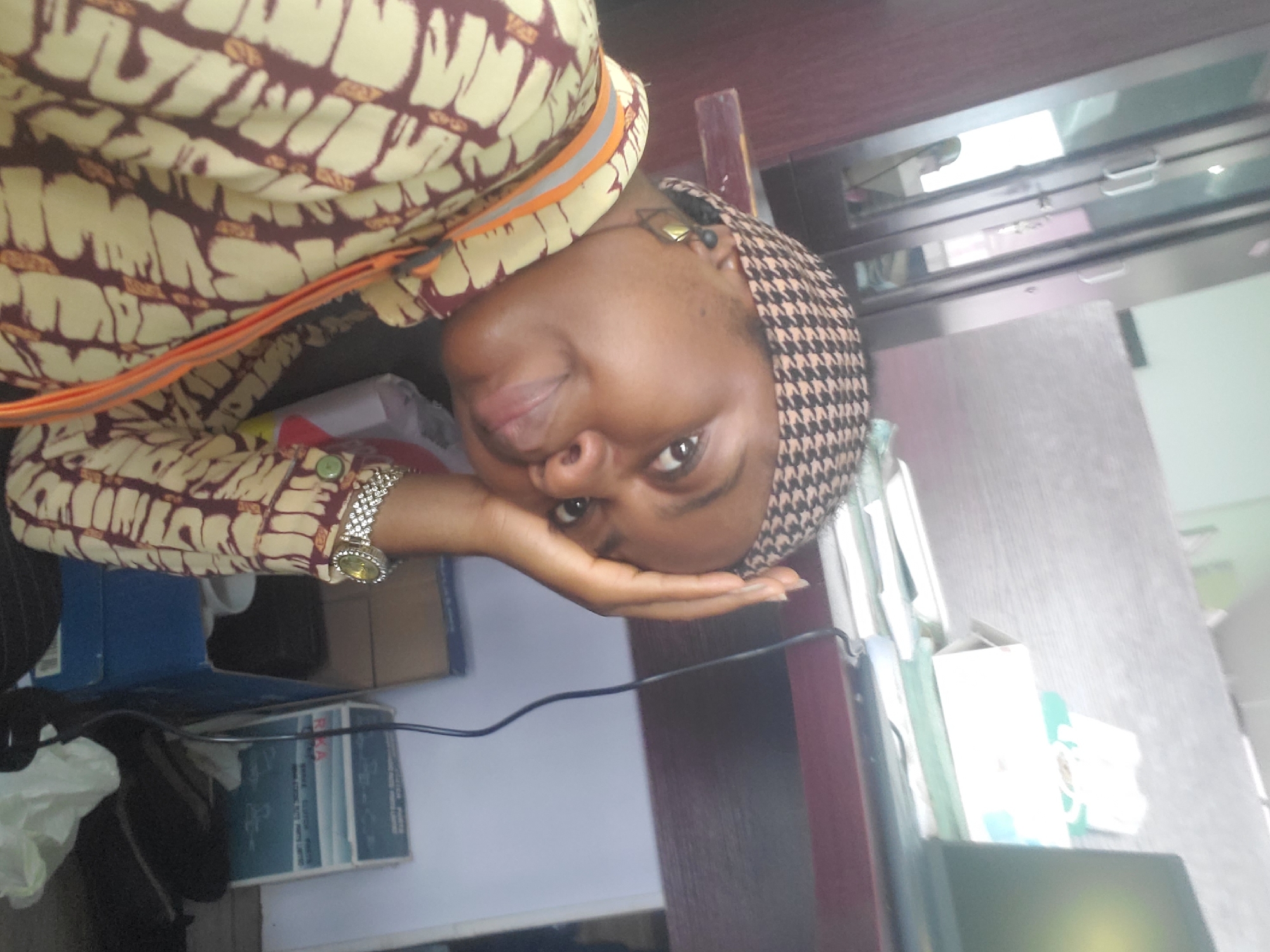 A medium-shot, eye-level, indoor, selfie captures a young Black woman in Gwagwada, Nigeria, leaning on her hand with her head tilted. She wears a patterned headscarf and a yellow and brown patterned long-sleeved shirt, accented with an orange and grey lanyard. A silver watch adorns her left wrist. The background reveals a desk with stacked papers, a laptop, and various boxes, suggesting an office or study environment.