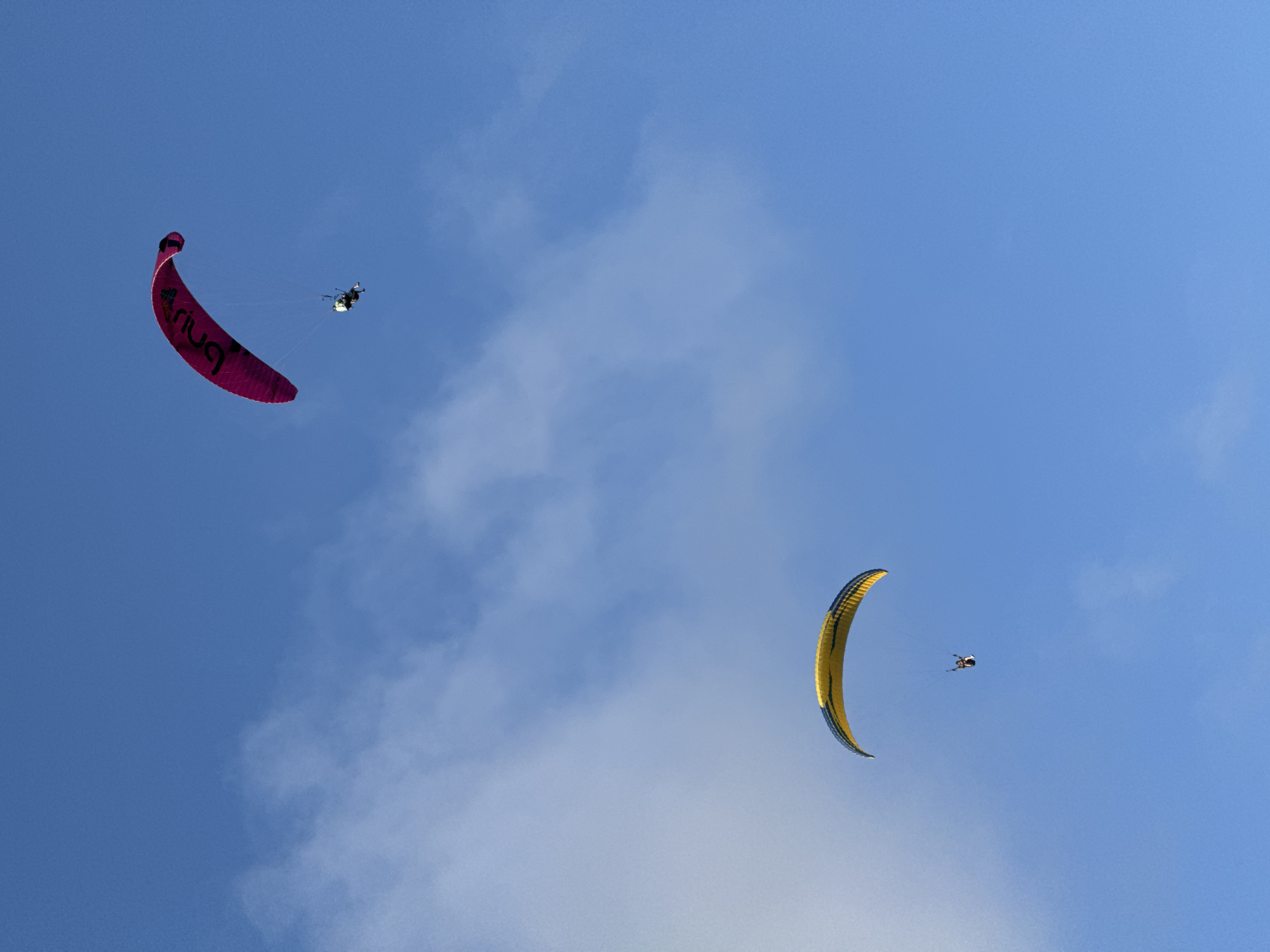 The photograph captures two powered paragliders against a backdrop of blue sky with scattered clouds. The paraglider in the upper-left has a pink canopy with the word "Ariua" printed on it in black, while the other paraglider is yellow with blue edges, located in the lower-right portion of the image. Each has a figure suspended below, presumably the paraglider pilot. The sky contains wisps of clouds, particularly in the lower-center and upper-left of the frame. This photograph was taken in Kuta Selatan, Indonesia.