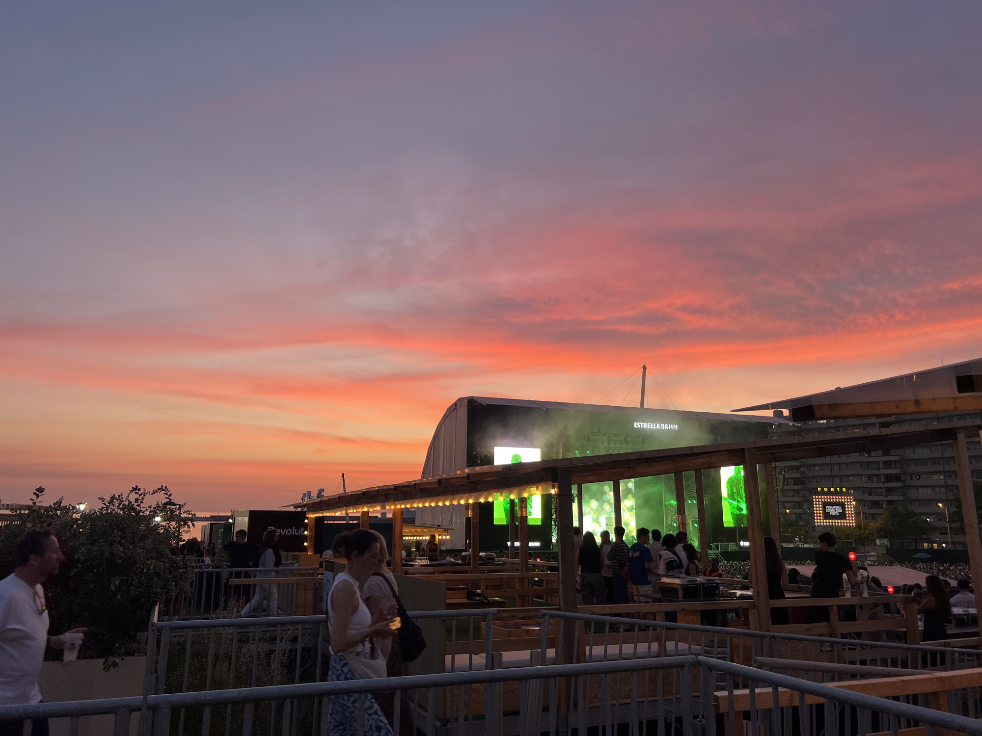 The image depicts an outdoor event at sunset in Porto, Portugal. A large crowd is visible in the background, gathered in front of a stage with a prominent "ESTRELLA DAMM" banner. In the foreground, people are milling around a raised viewing area with wooden structures and railings. The sky is a vibrant mix of orange, pink, and purple hues, characteristic of a sunset. The overall atmosphere suggests a music festival or concert.