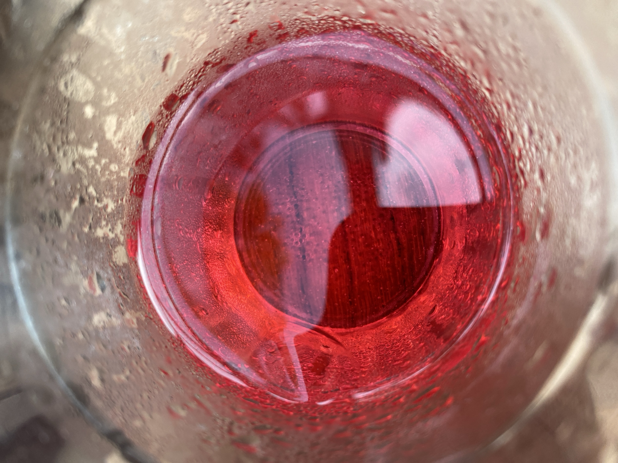 A close-up, overhead shot shows the surface of a red liquid in a clear glass. Condensation droplets are visible on the exterior of the glass. The liquid appears to be smooth, with slight ripples or undulations on its surface. The lighting creates reflections and highlights on the liquid and glass. The background is blurred and indistinct, suggesting an indoor setting. There are no people or specific landmarks visible.