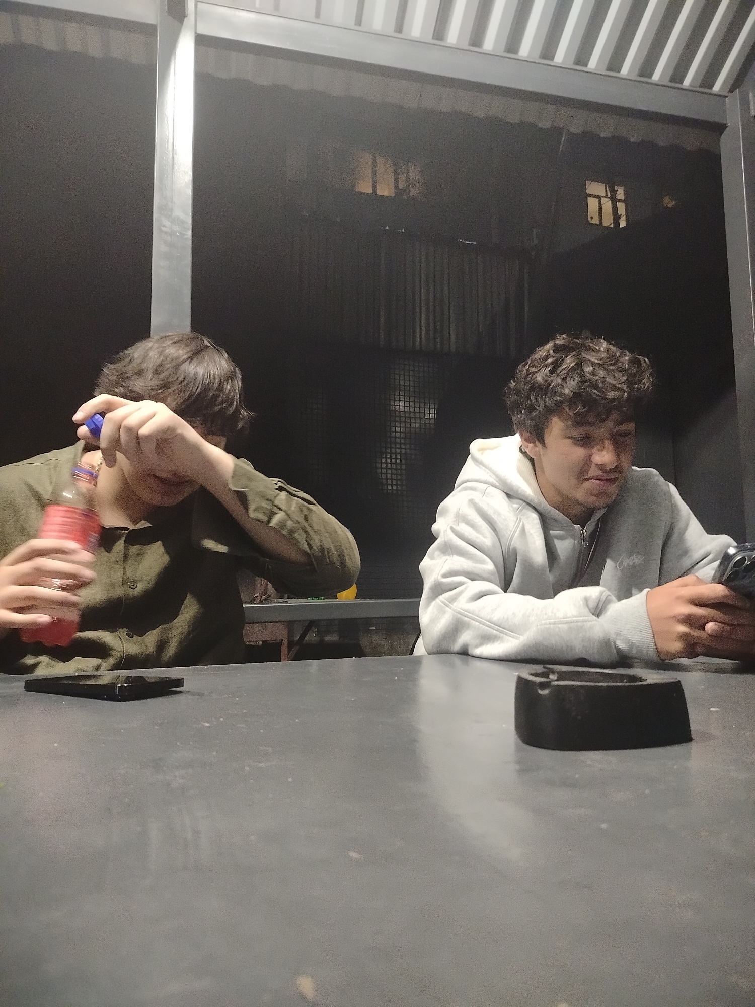 Two young men sit at a gray table under a partially covered outdoor structure. The man on the left wears a green button-up shirt and holds a bottle of pink liquid. His face is partially obscured by his hand. A dark object, possibly a phone, rests on the table to his left. The man on the right wears a gray hooded sweatshirt and looks at a mobile phone in his hands. An ashtray sits on the table in front of him. In the background, through the structure, a building with a lit window is visible. Given the setting, the location might be an outdoor cafe or restaurant. Yerevan, Armenia is not confirmed; location cannot be confirmed by image alone.