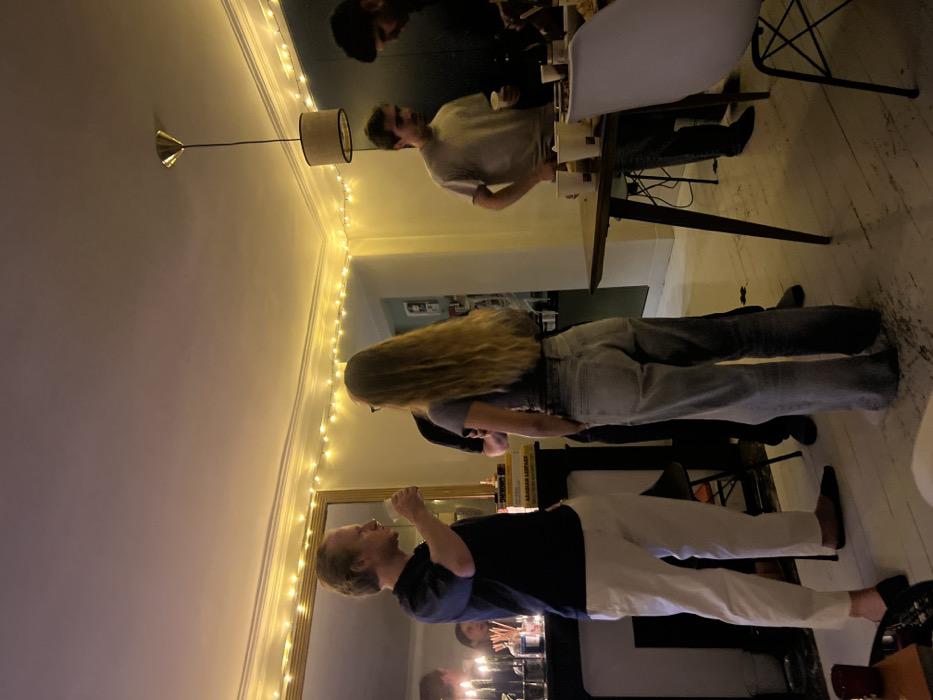 A sideways indoor shot reveals a gathering of people. A man in a navy shirt and white pants is on the left side. A woman with long blonde hair is beside him. Further back, two men are around a table with drinks. String lights decorate the ceiling near a lamp. There's a mirror and a glimpse of a painting or picture frame in the background. The floor appears to be wooden.