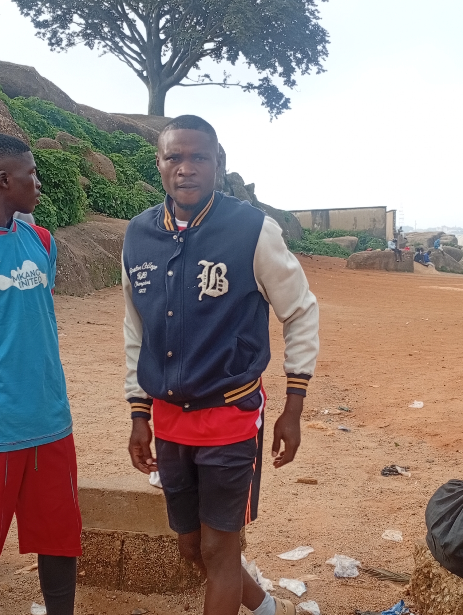 The image depicts a street scene in Jos, Nigeria, on a cloudy day. The primary subject is a young Black male standing in the foreground, facing the camera. He is wearing a navy blue and white letterman jacket over a red t-shirt and dark shorts. The jacket has yellow and navy blue stripes on the cuffs and collar, and features an embroidered "B" emblem on the right breast and text on the left breast that reads "Boston College 89 Champions 1972".
To the left of the main subject, another young Black male is partially visible, facing away from the camera. He is wearing a light blue t-shirt with white lettering that reads "MKANG UNITED" and red shorts.
The background reveals a natural landscape with large, weathered rocks and lush green vegetation. A large tree with spreading branches stands prominently on a rocky outcrop. In the mid-ground, there are several other individuals, some seated on the rocks, and structures that appear to be buildings or walls. The ground in the foreground is sandy and littered with various debris, including plastic bags and wrappers. The overall impression is that of an outdoor gathering or recreational area in Jos.
