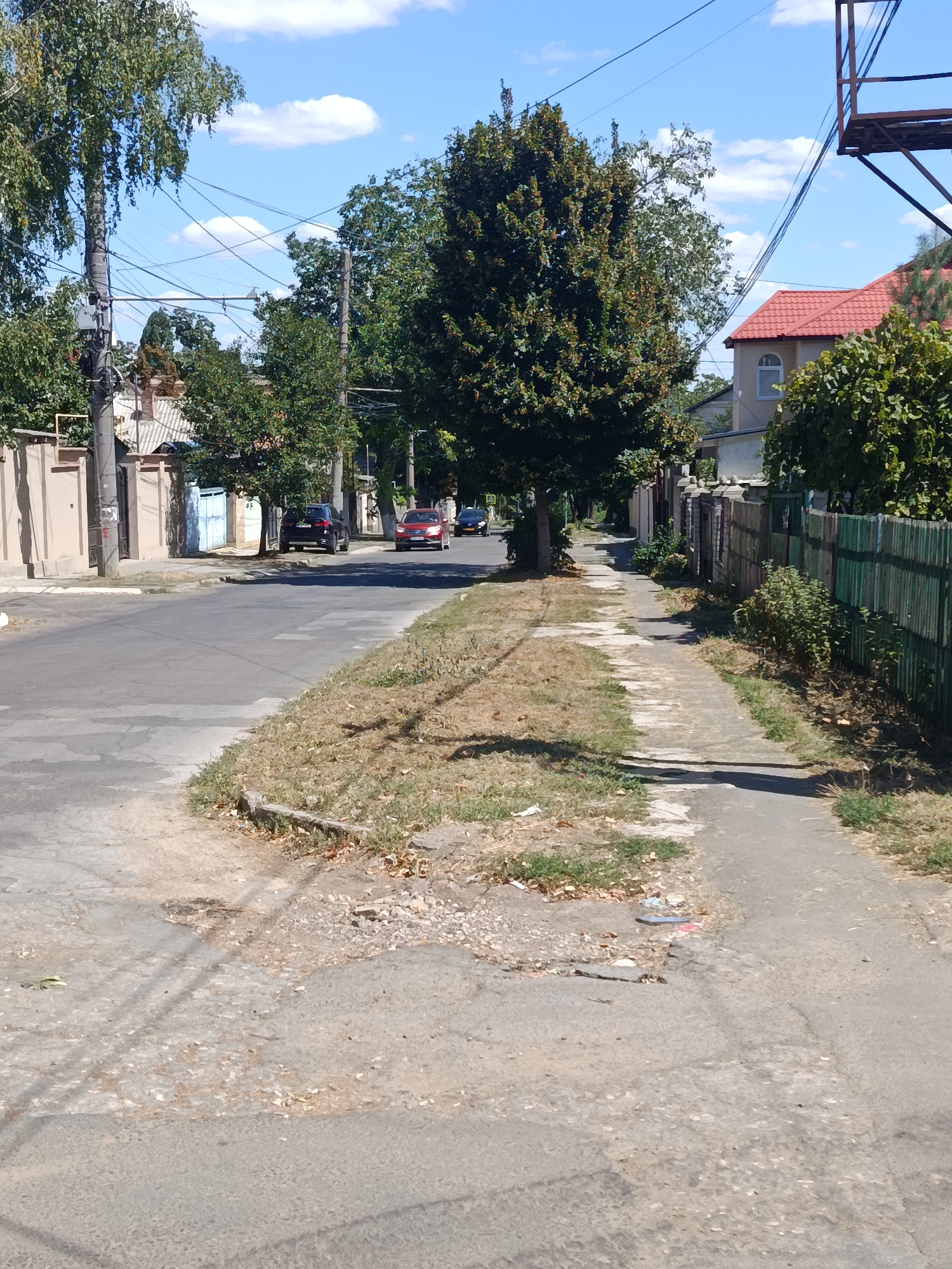 The image depicts a street scene in Chișinău, Moldova, on a sunny day. A paved road runs horizontally across the lower portion of the frame, with a concrete sidewalk on the right side. The sidewalk is uneven and cracked in places, with dry grass and some debris. Several trees with green foliage line the street and sidewalk.
On the road, there are a few parked cars, including a black SUV and a red sedan. The traffic appears light. The buildings on either side of the street are mostly residential, with fences and walls visible. A prominent feature is the dense network of overhead electrical wires crisscrossing the sky, supported by utility poles. The sky is blue with scattered white clouds. The overall impression is that of a typical residential street in a city.