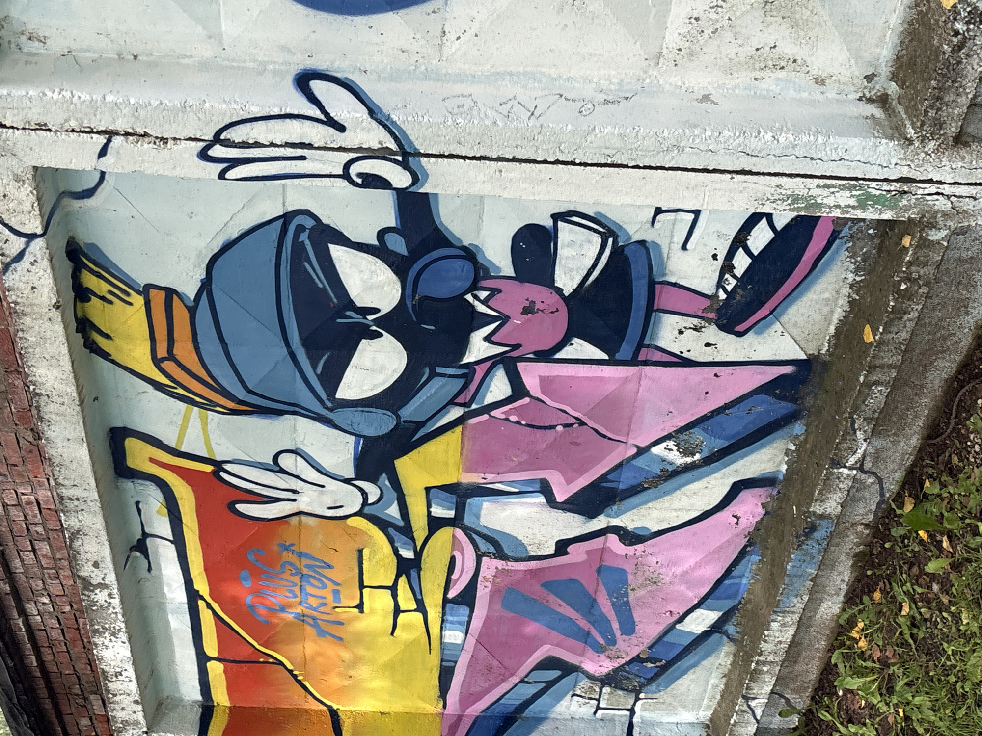 The image depicts graffiti art on a concrete wall. The graffiti features a cartoon character with a blue helmet and a pink dress, along with abstract shapes in pink and blue. The character has large white eyes with black pupils and is raising its hands, with one hand in a peace sign. Behind the character, there are abstract shapes in shades of red, yellow, and orange, and also in pink and blue. The wall is weathered and cracked, with some graffiti appearing faded or chipped. At the top of the wall, there is a brick structure. The base of the wall shows signs of dirt and vegetation, with some leaves on the ground. The location is identified as Krasnokamsk, Russia.