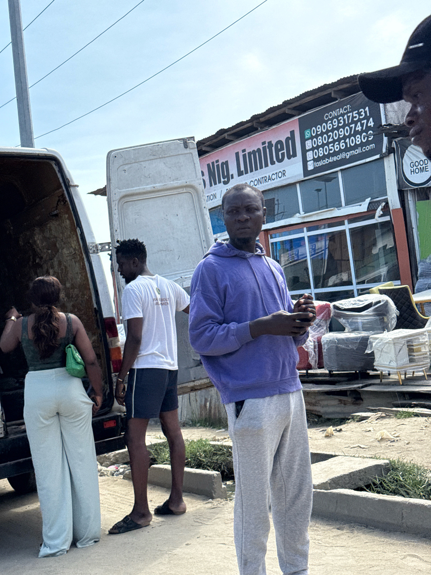 The image depicts an outdoor scene in Ado, Nigeria. In the foreground, a young Black man in a purple hoodie and grey sweatpants stands facing the camera with his hands clasped. To his left, a Black man in a white t-shirt and dark shorts is looking away from the camera, near the open back of a white van. A Black woman wearing a green sleeveless top and light-colored wide-leg pants is also near the van, facing away from the camera and looking into it. Behind the people, a storefront displays furniture, with a sign that reads "Nig. Limited" and contact information. The environment appears to be a commercial area, with a dusty ground and visible infrastructure like a utility pole and overhead wires. The overall impression is of a transactional or logistical activity related to the furniture business.