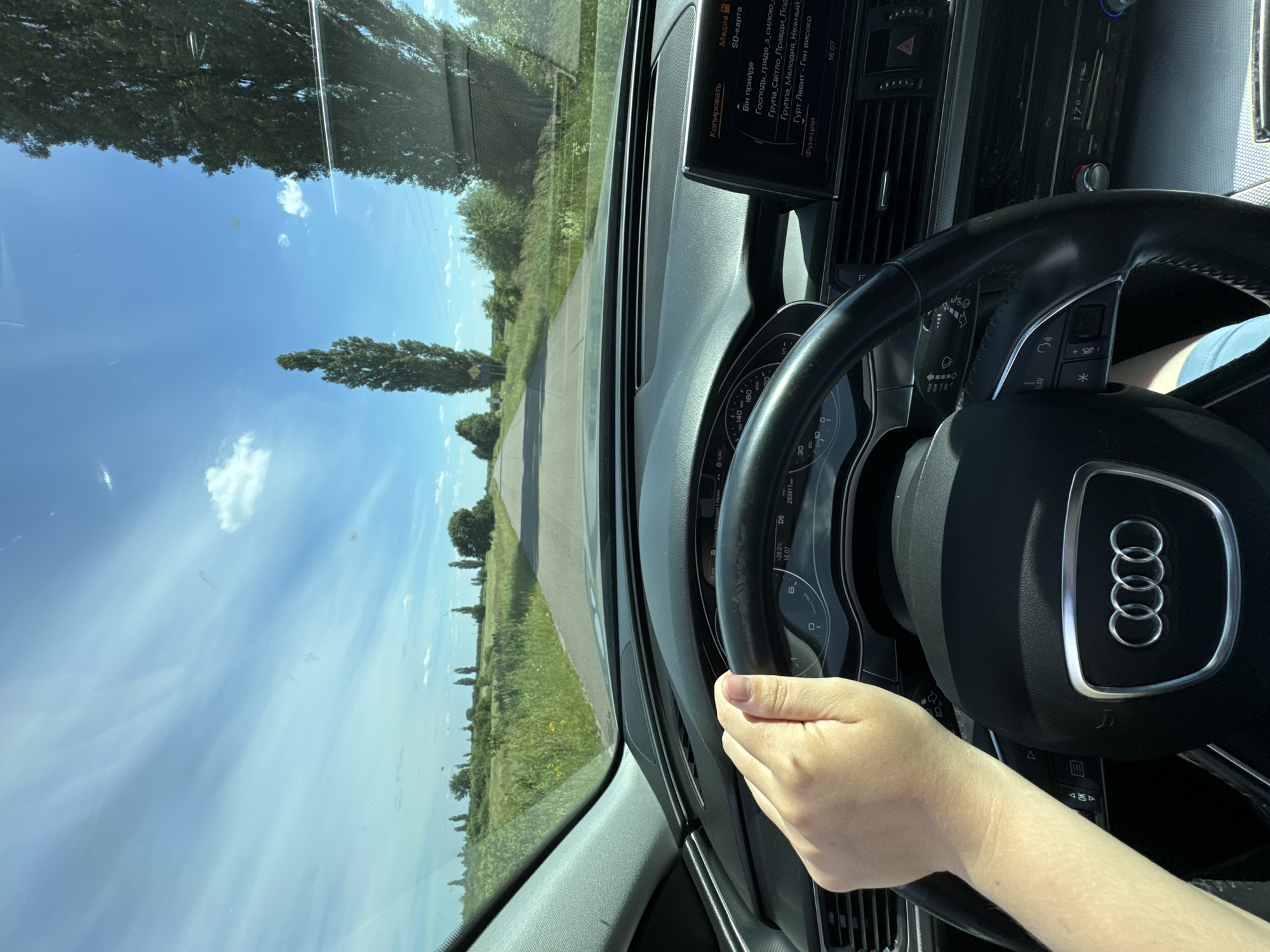 The image captures a first-person perspective from the driver's seat of a car, likely an Audi, as it travels along a paved road through a rural landscape. The driver's left hand is visible on the steering wheel, a portion of the dashboard with a digital display and climate controls are also in view. The road ahead is straight, bordered by lush green fields and tall, slender trees on either side. The sky above is a bright blue with scattered white clouds, indicating a sunny day. The digital display on the dashboard shows navigational information and media playback details, including song titles in Ukrainian. The scene suggests a journey through the countryside, possibly near the village of Dovhe, Ukraine, given the car's navigation system and the characteristic landscape.