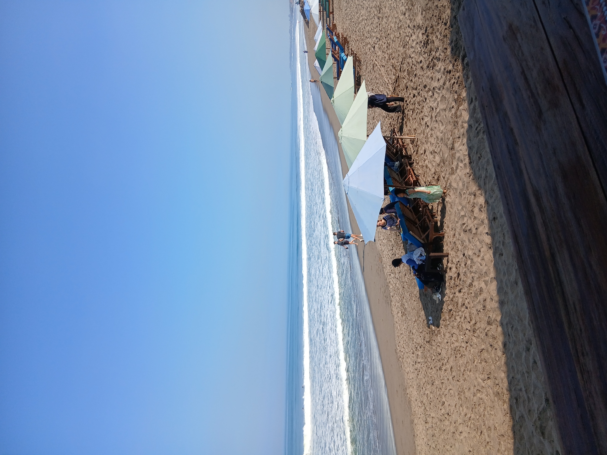 This image captures a serene beach scene in North Kuta, Indonesia, under a clear blue sky. The foreground features a wooden surface, possibly a table or deck, with a textured, dark brown finish. Beyond this, the sandy beach stretches out, populated by several lounge chairs and umbrellas, indicating a resort or public beach area.
A row of umbrellas, in shades of white, light blue, and various greens, provides shade for the lounge chairs. Several people are visible: a woman in a light green dress stands near a white umbrella, a person in dark clothing is standing near a row of chairs, and a couple is walking along the shoreline in the middle ground. In the background, the ocean meets the sand, with gentle waves breaking and white foam creating a distinct line. A few surfers can be seen in the distance on the water. The overall ambiance is one of a peaceful day at the beach, with a mix of leisure and natural beauty.