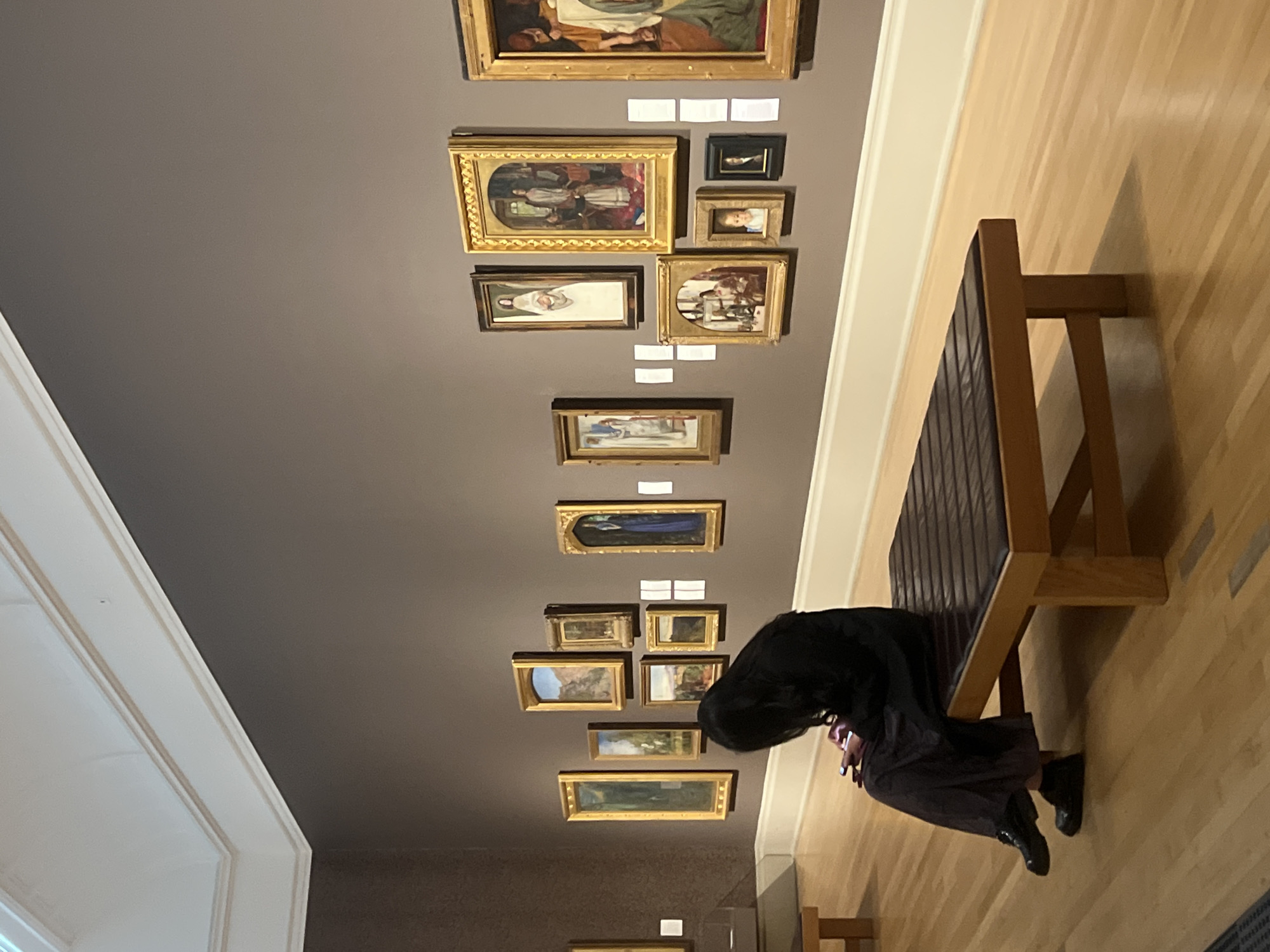 This image captures an interior scene within an art gallery or museum, likely located in London, United Kingdom. The primary subjects include a person and numerous framed artworks displayed on a wall.
The central focus is a dark grey wall adorned with a collection of paintings, varying in size and orientation. Many of the artworks feature ornate gold frames, while others have simpler dark frames. The paintings depict a range of subjects, including portraits, figure studies, landscapes, and possibly still life. Small, rectangular white labels are visible beneath several of the paintings, providing exhibition information, though the text on them is not legible.
In the lower right portion of the image, a person is seated on a long, dark brown upholstered bench with a light wooden frame. The person has long, dark hair and is dressed in dark clothing. They are looking down at a mobile phone, which emits a soft light onto their face, suggesting they are actively using the device.
The floor is made of light-colored wooden planks, complementing the gallery aesthetic. Along the top edge of the wall, there is a white architectural trim or crown molding, indicating the transition to a ceiling or upper wall section. The overall lighting appears artificial and even, typical of an indoor exhibition space. No specific time of day or weather conditions are discernible, as the scene is indoors. The activity taking place is generally viewing art, though the individual shown is momentarily engaged with their phone.