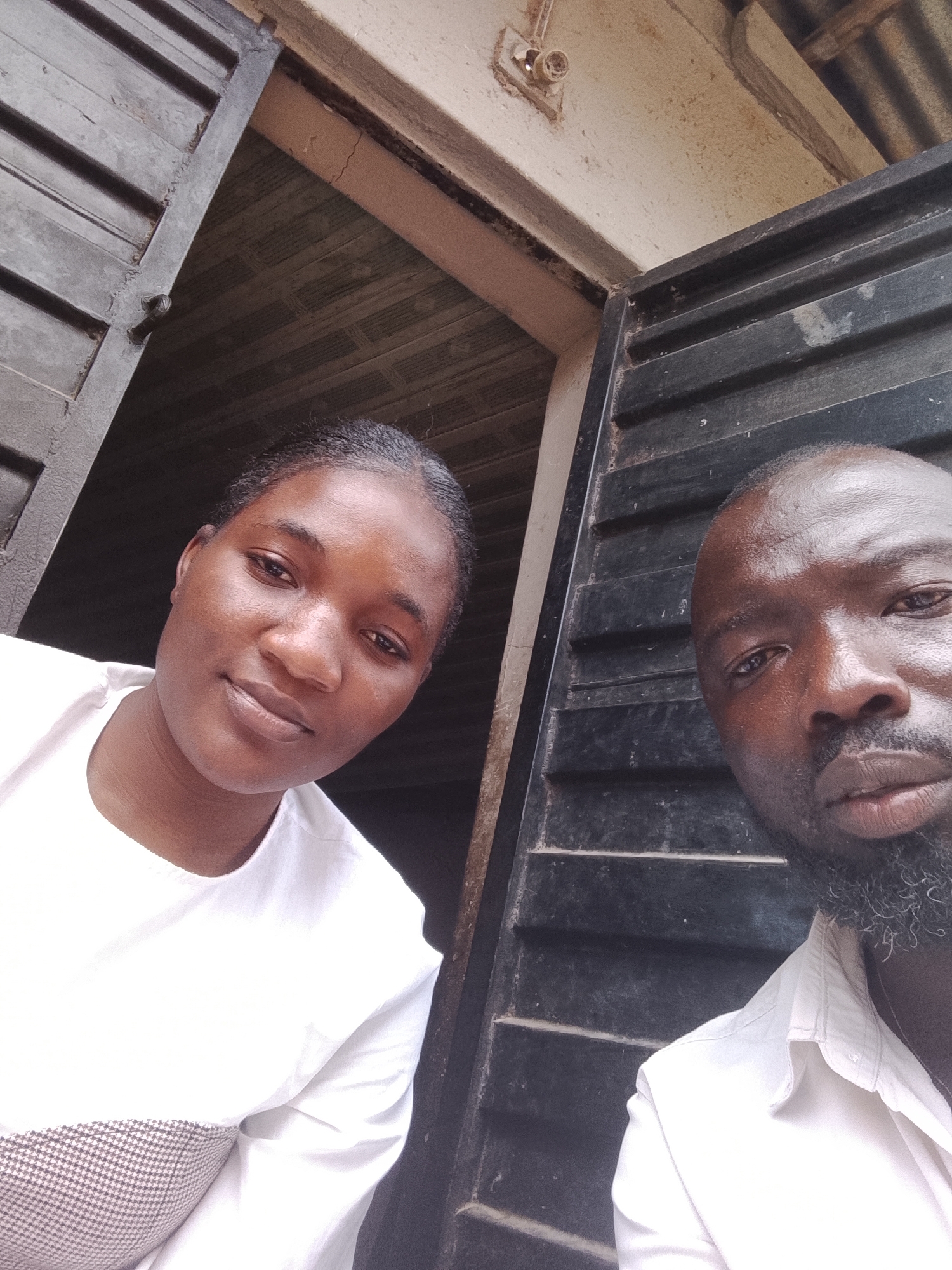 Two individuals are positioned close to each other in an indoor setting. One is a young woman wearing a white shirt. Beside her, a man with a beard is also wearing a white shirt. They are both looking at the camera. Behind them is a dark window or door. An electrical fixture is visible in the upper part of the image. The location is possibly within the city of Jos, Nigeria.