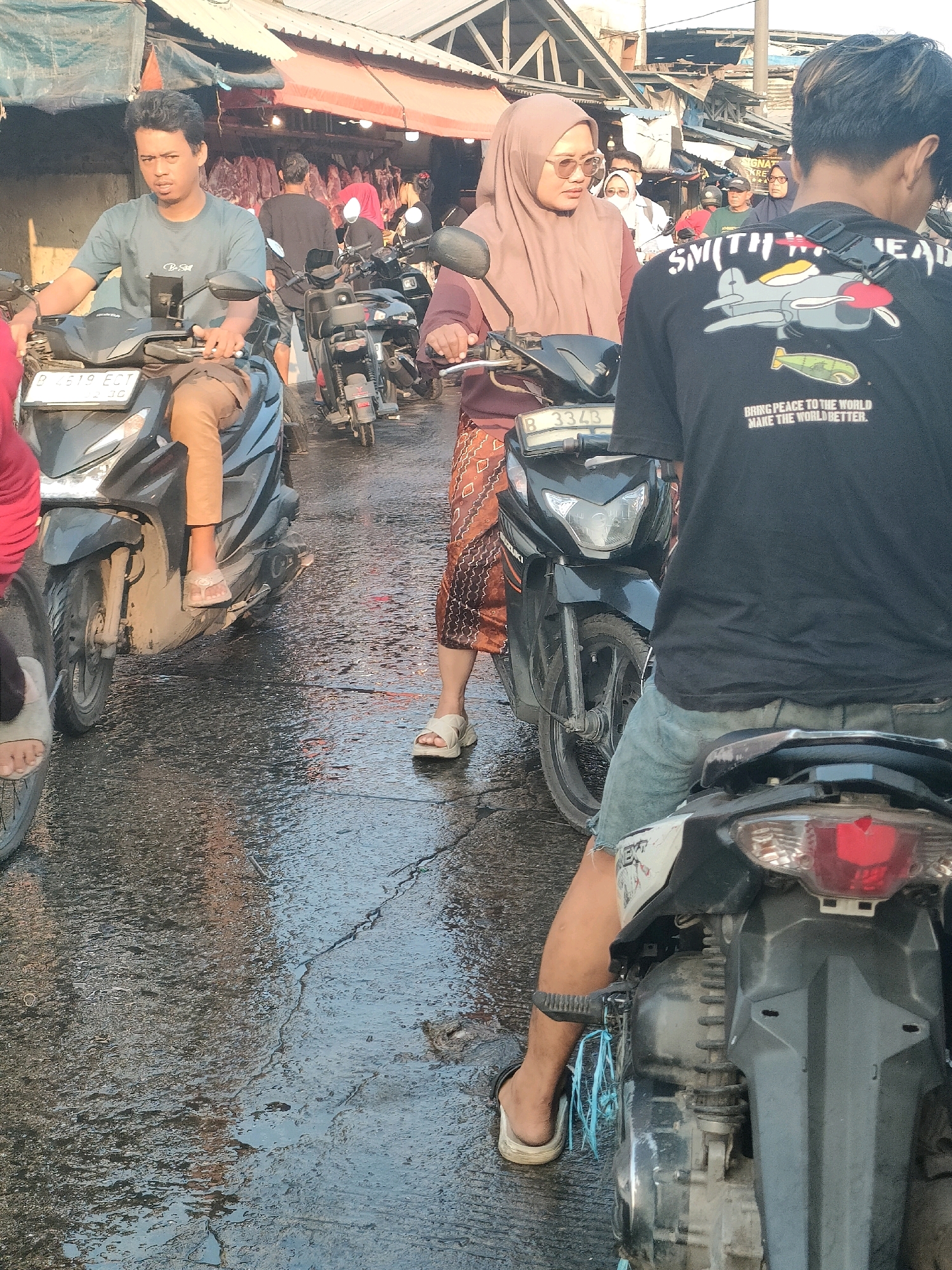 In a street market scene in Bekasi, Indonesia, a man in a teal t-shirt rides a black scooter, looking forward. Behind him, a woman in a taupe hijab and sunglasses rides another black scooter, also looking forward. A third individual wearing a black t-shirt with a graphic print of an airplane and text is seated on a motorcycle in the foreground, facing away from the camera. The ground is wet, suggesting recent rain or market activity. Several other motorcycles are visible, along with market stalls and awnings in the background, indicative of a bustling commercial area.