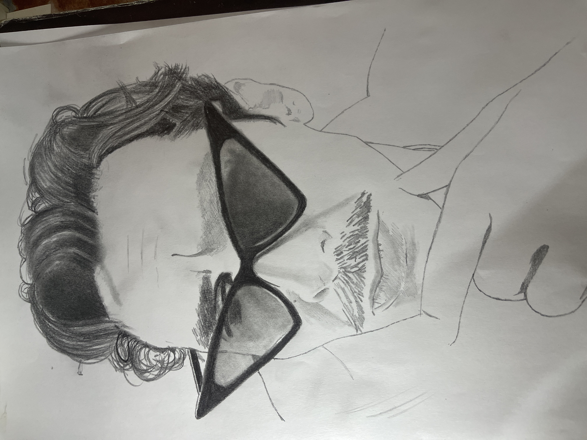 The image is a pencil drawing of a man with dark hair, wearing sunglasses. The drawing is on a white piece of paper. The man is lying on his side, possibly on a bed or soft surface. The focus is on his head and shoulders. No contextual clues suggest the location of Jos, Nigeria.