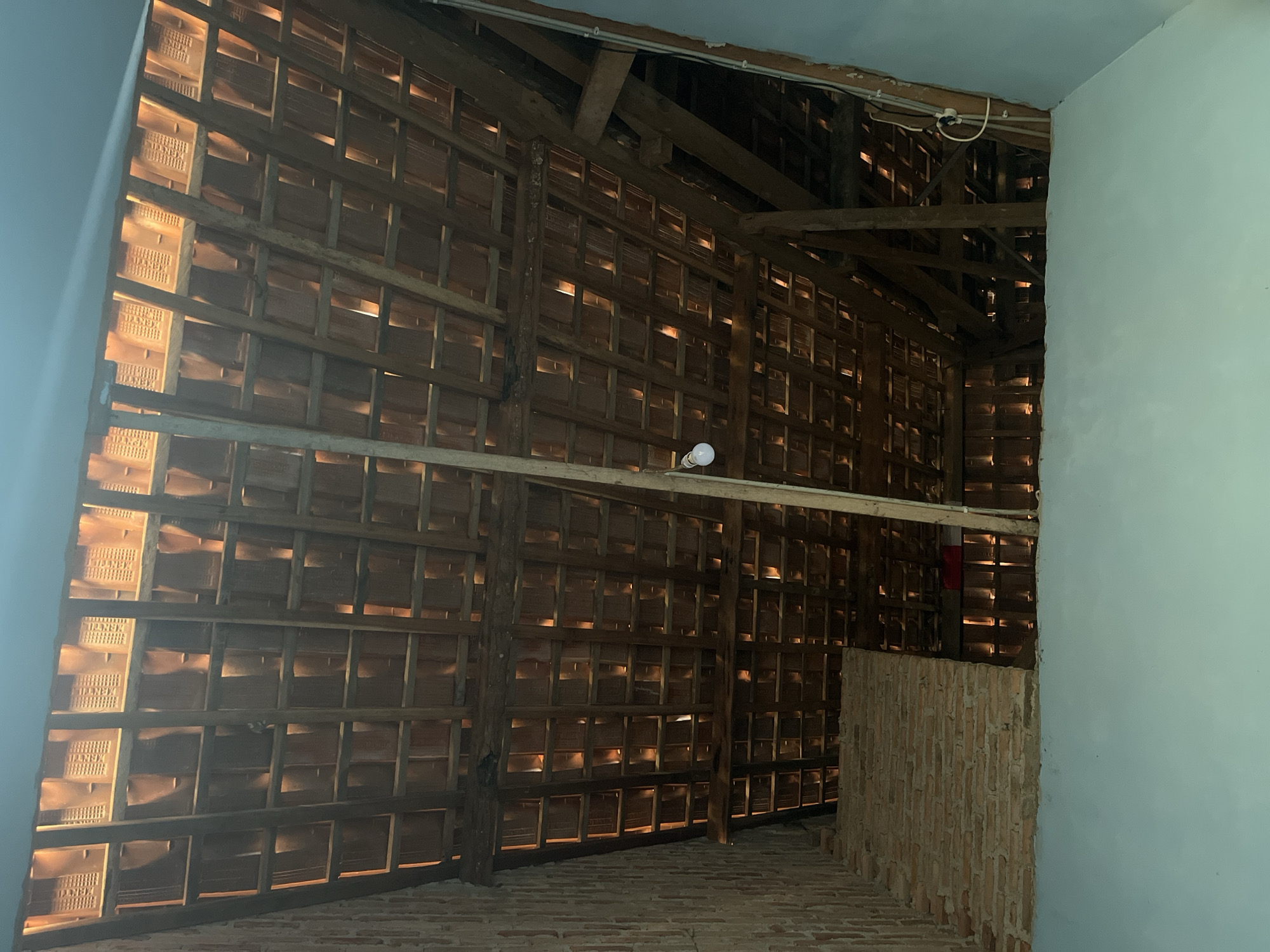The image depicts the underside of a tiled roof structure, likely an attic or unfinished room, in Kebumen, Indonesia. The roof is constructed with wooden beams and rafters supporting rows of traditional terracotta roof tiles. Light filters through gaps between the tiles, creating a pattern of illuminated and shadowed areas. A brick wall is visible on the left side, and a lightbulb hangs from a wire in the upper center of the frame. The overall impression is one of an unfinished or rustic interior space.
