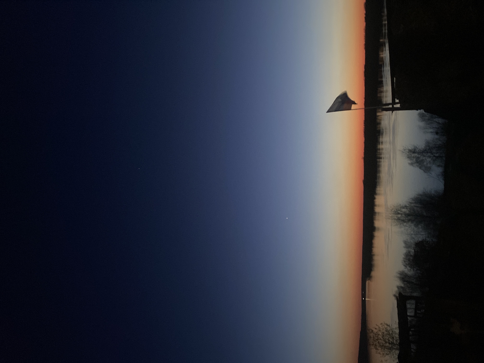 The image is a horizontal view of a sunset scene. The sky transitions from dark blue on the left to orange and light blue towards the right, indicating the setting sun. A body of water reflects the sky's colors. On the right, the silhouette of trees and vegetation lines the bank. A flag is visible on the bank near the center, its shape indicating a breeze. The location is likely Ольховка, Russia, suggested by the environment and the flag.