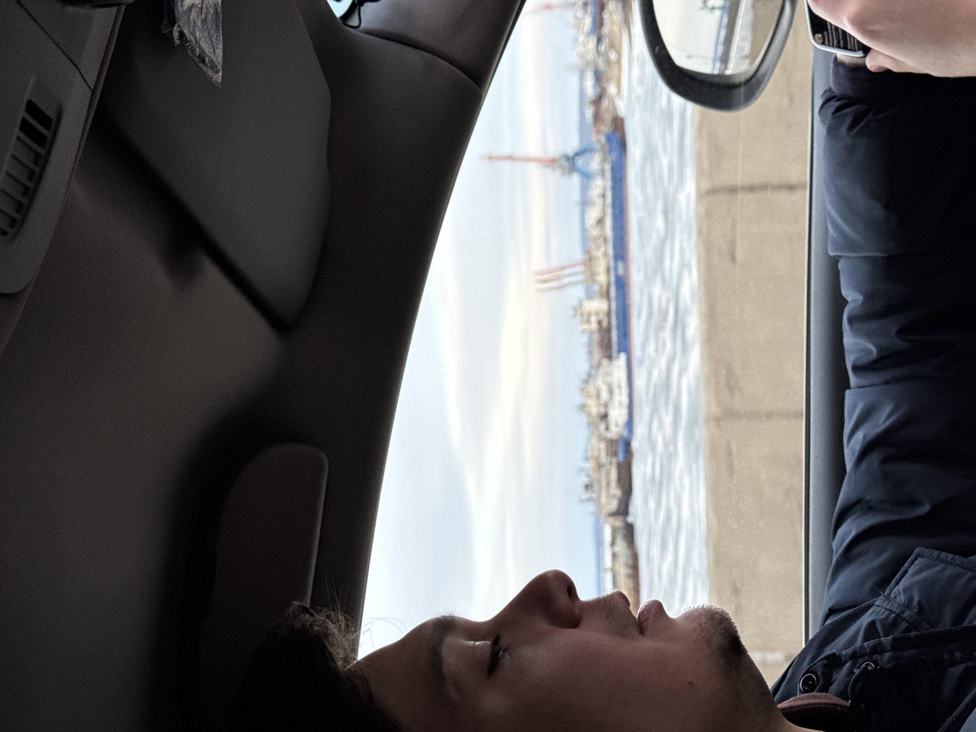 A man is seen from the chest up, lying down in what appears to be the passenger seat of a car. The car's interior is visible, including the dashboard and the passenger side window. Outside the window, a body of water and a skyline with what appears to be industrial cranes can be seen. The scene is Chaikovsky, Russia.