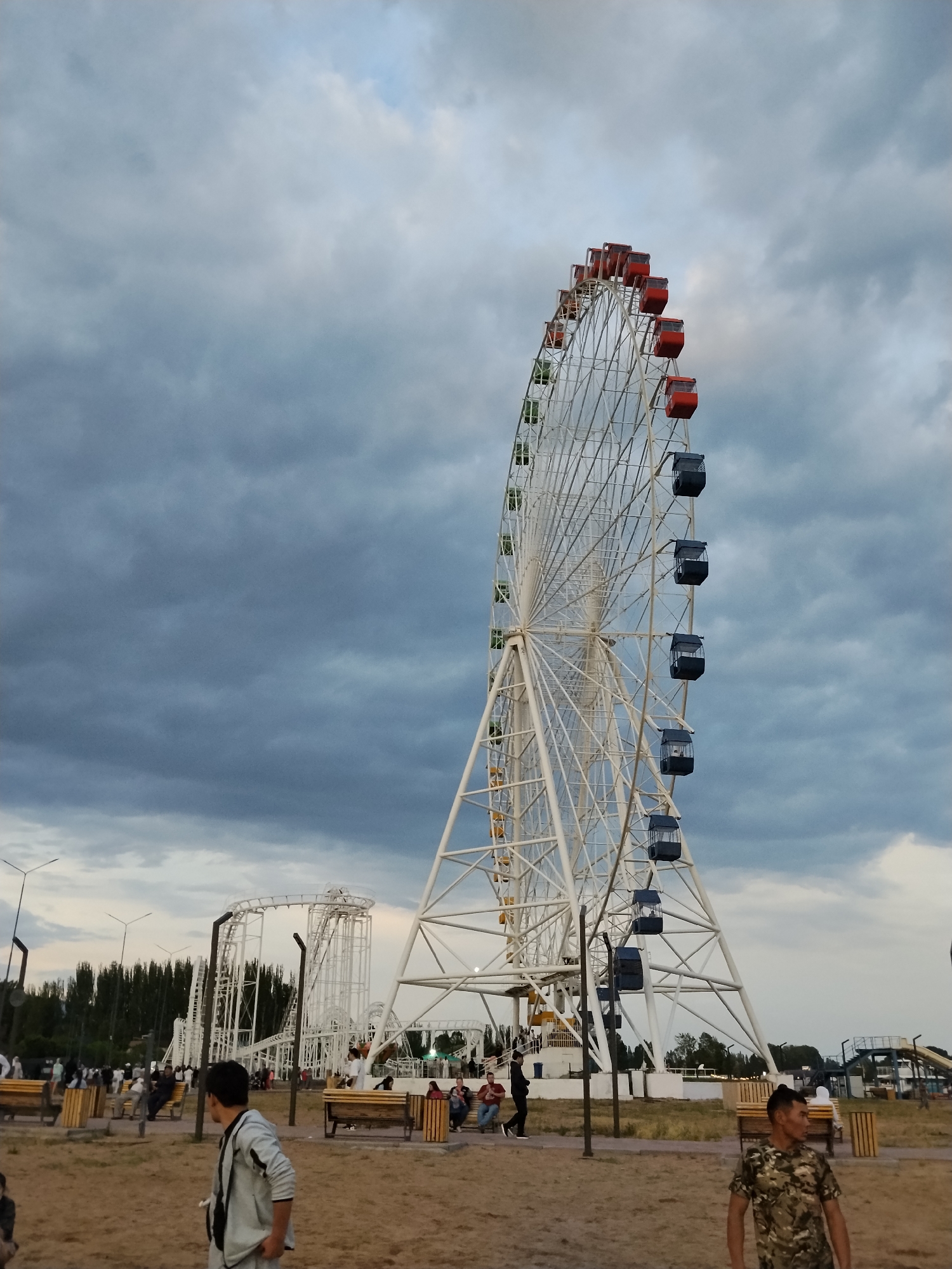 An outdoor, eye-level, wide shot captures a large Ferris wheel with red and dark blue gondolas against a cloudy, gray sky. The Ferris wheel is white and has a complex lattice structure. To the left of the Ferris wheel, a white roller coaster track is visible. In the foreground, several people are scattered throughout a park-like setting. Two men are walking in the foreground, one wearing a gray jacket and the other a camouflage t-shirt. Other individuals are seated on benches or walking in the midground. The ground is covered in dry grass and sand, and trees line the background.
