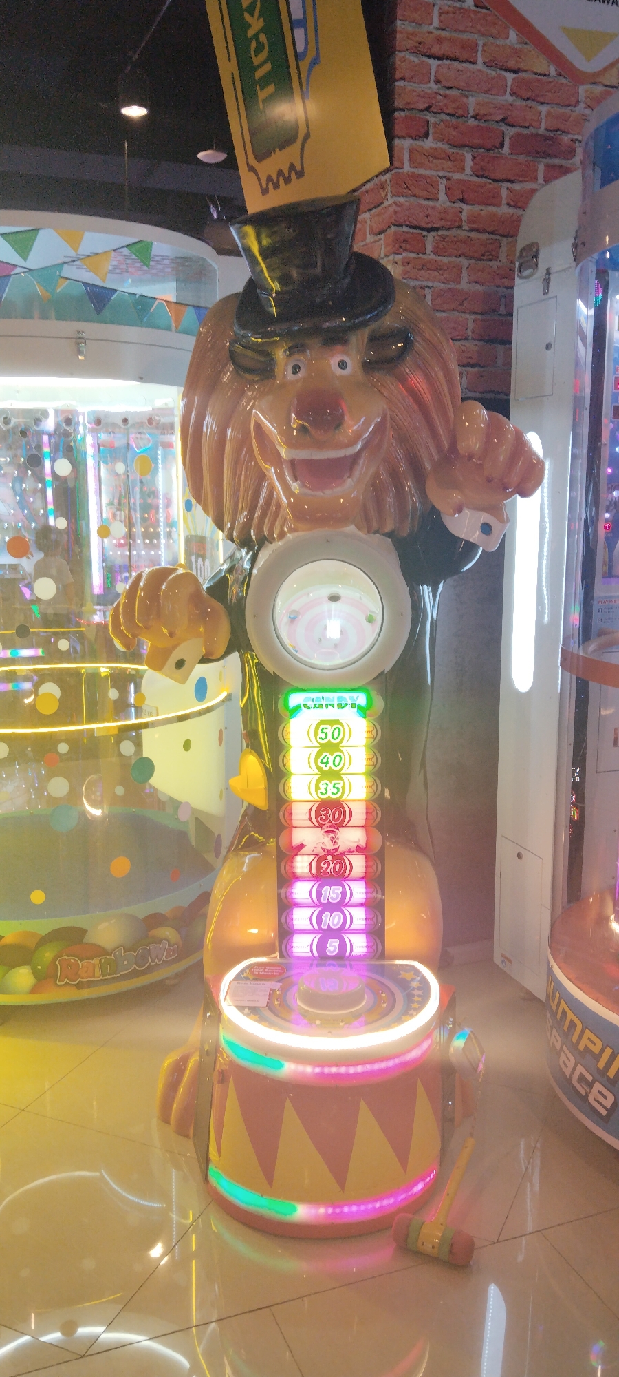 This is a medium shot, eye-level, full-frame photograph of an arcade game machine, likely located in Semarang, Indonesia. The machine features a large, anthropomorphic lion character wearing a black top hat and a green jacket. The lion has a wide, toothy grin, with its eyes looking slightly upward and to its right. Its right hand is extended towards the viewer, while its left hand is holding the brim of its hat.
Below the lion's head is a circular screen with colorful lights and what appears to be a spinning element inside. Beneath this screen is a vertical display with a list of numbers: 50, 40, 35, 30, 20, 15, 10, and 5, each enclosed in colored segments that transition from yellow and green at the top to pink and purple at the bottom. The word "CANDY" is illuminated in green above the numbers.
The lion character is positioned in front of a large, clear cylindrical display filled with colorful round objects, possibly candies or balls, and the word "Rainbow" is visible on the base of this display. To the right of the lion machine, a portion of another arcade game with the word "JUMPING" visible is present.
The floor is shiny and reflective, with a pattern of light and dark tiles. A colorful, drum-like base with illuminated bands of pink, yellow, and green lights supports the main lion character. A small, yellow and pink toy hammer rests on the floor in front of this drum base. The background features a brick-patterned wall and what appears to be other arcade game machines and displays, all illuminated with bright, colorful lights.