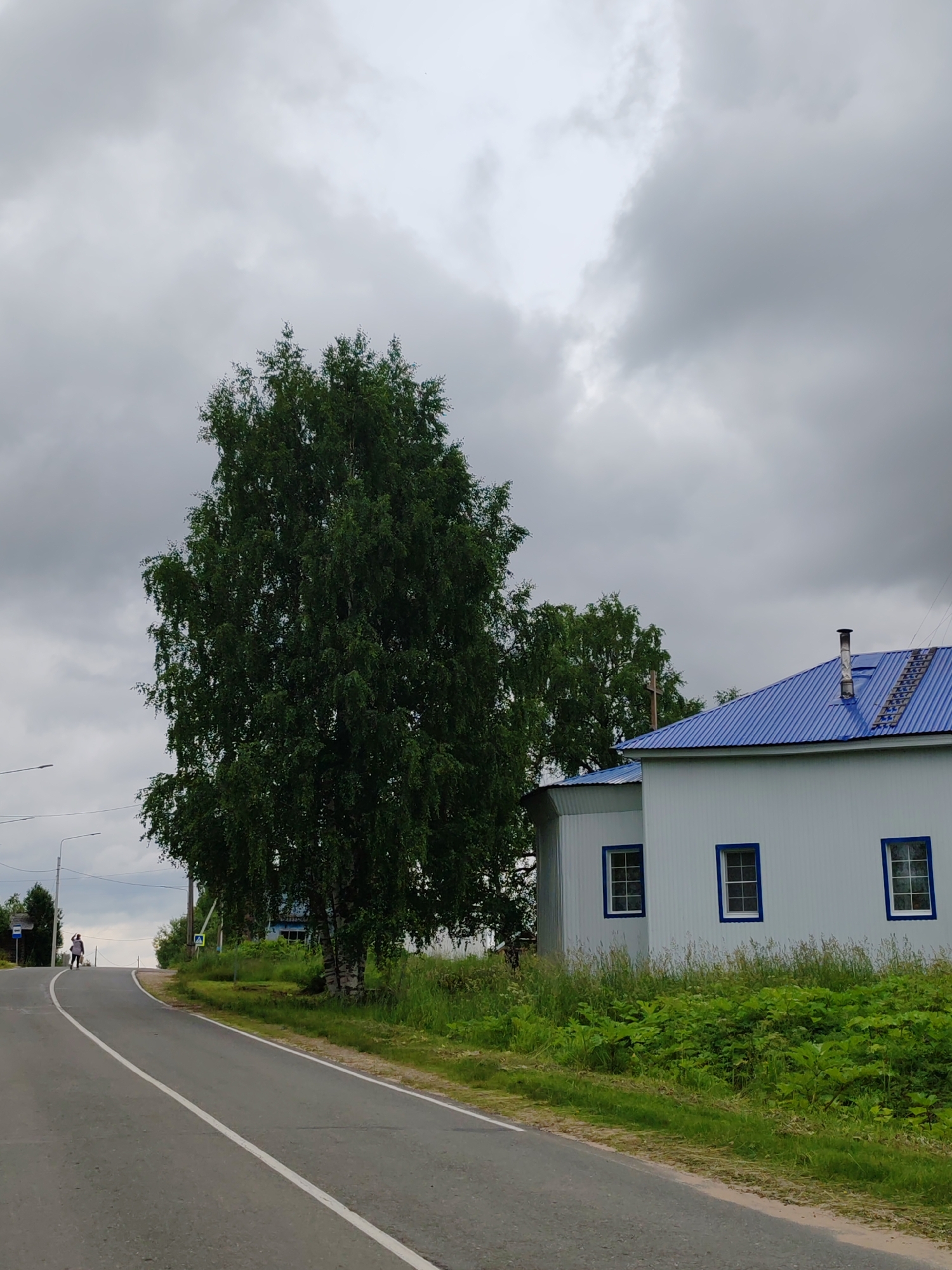 The image depicts a rural road curving uphill under a cloudy sky. A tall, leafy birch tree stands prominently on the left side of the road. To the right, a white building with a blue metal roof and blue-trimmed windows is visible. There is lush green vegetation along the roadside. In the distance on the left, a person is walking up the road. The location is identified as the city of Chukhlem, Russia.