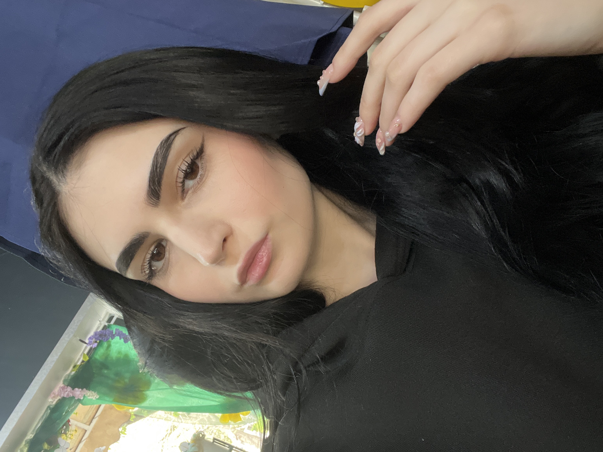 A close-up portrait of a woman with long, dark hair, light skin, and brown eyes. She is wearing makeup, including eyeliner, blush, and lipstick. Her nails are long and decorated with a white and silver design. She is wearing a black top. Behind her, a blue garment hangs. A window is also visible, decorated with colorful flowers and greenery. The location in Burgas, Bulgaria cannot be verified from this image alone.