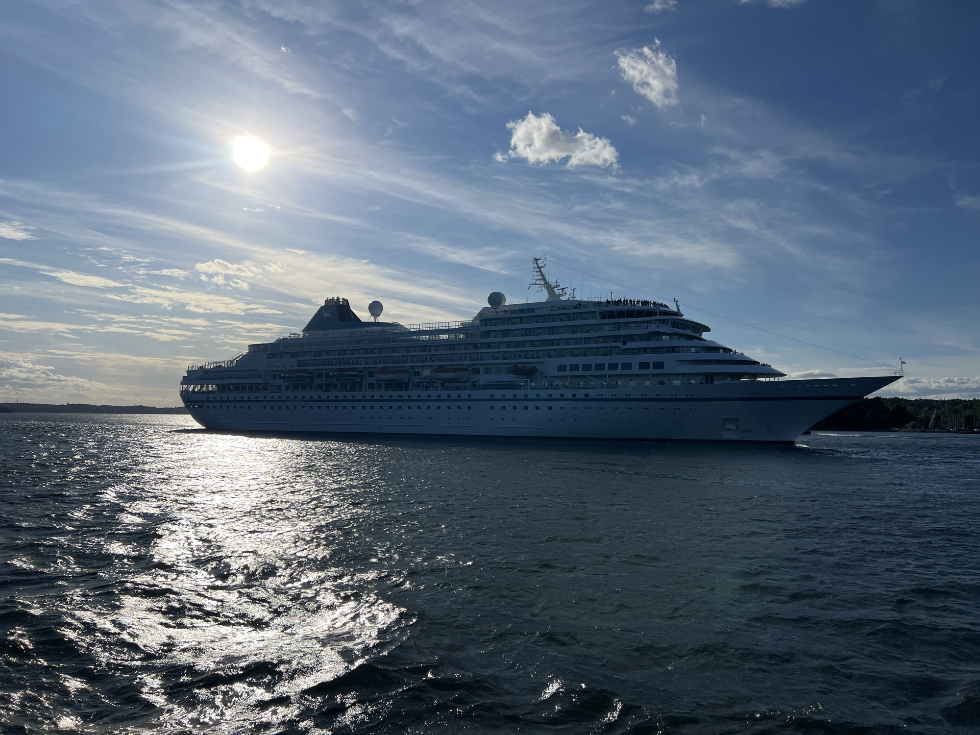 A cruise ship, identified by the name "AMPHORA" on its bow, sails on dark blue water under a bright sun and a sky with scattered white clouds and wispy cirrus formations. The sun's rays create a strong glare on the water's surface. The ship is large, white, with multiple decks, and a dark green funnel at the stern. People are visible on the upper decks of the ship. The shoreline, with trees and some structures, is visible in the background to the right. The location is the city of Boo, Sweden.