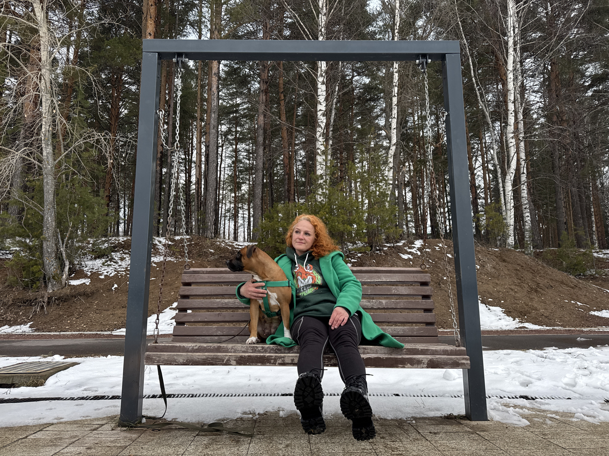 A woman with curly red hair sits on a swing bench with a brown dog wearing a green harness. She is wearing black pants, black boots, and a green coat over a green hoodie with a graphic design. The swing bench has a gray metal frame and wooden slats. The background is a forest of tall, thin trees, including birch trees with white bark. The ground is covered with a patchwork of snow and exposed ground. The location is Chaikovsky, Russia. The woman is holding the dog.