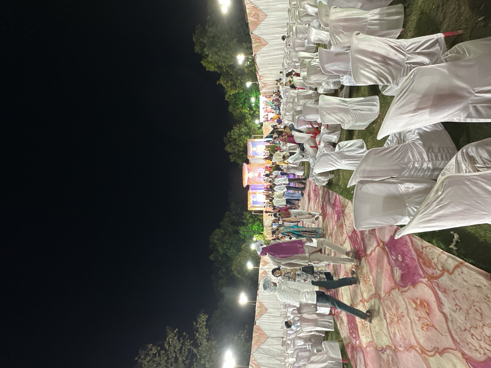 The image depicts an outdoor event in Shahapur, India, at night. Rows of white chair-covered chairs are visible under a large tent, indicating a formal gathering. People are present, with some walking on a patterned ground covering. The area is lit by overhead lights. The background features trees and a dark sky.
