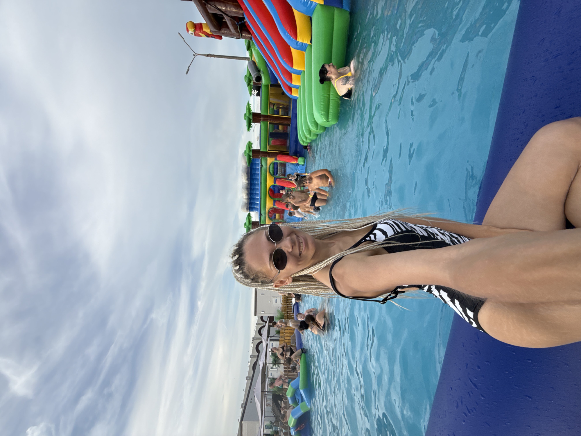 The image depicts a lively scene at an outdoor water park, likely in Rusanovo, Russia. The main subject is a young woman with blonde braided hair, wearing a black and white striped bikini and sunglasses. She is smiling and taking a selfie while posing in a blue swimming pool.
In the background, several other people are visible in the water and around inflatable water slides. One prominent inflatable structure features bright red, blue, and green sections, resembling a pirate ship with slides and climbing areas. Some individuals are partially submerged in the water, while others appear to be interacting with the inflatable attractions. The water is a clear, bright blue, and the sky above is partly cloudy, suggesting it is daytime.
The overall atmosphere appears to be one of fun and recreation, with people enjoying a day at the water park. The presence of umbrellas and some building structures in the background hints at a developed recreational facility. There is no visible text in the image.