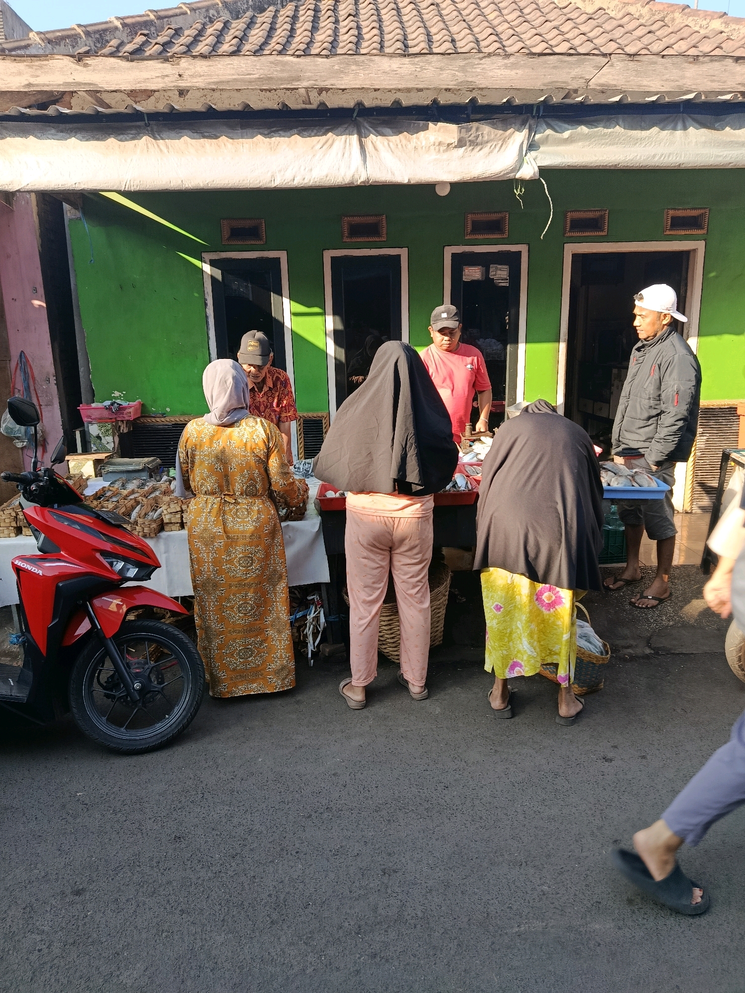 In Gondanglegi, Indonesia, a street market scene unfolds under a tiled roof structure. A red Honda motorcycle is parked on the left. Several individuals are engaged in transactions at a market stall. Two women, one in a patterned orange dress and the other in a yellow floral skirt and black head covering, are facing away from the camera, interacting with a vendor. The vendor, a man in a red shirt and black cap, stands behind the stall, which displays fish on blue trays and other goods in baskets. Another man, wearing a black jacket and a white cap, stands to the right, observing the scene. The backdrop is a bright green building with multiple entrances. The ground is paved with asphalt. The lighting suggests it is daytime, with shadows indicating the sun is at a moderate angle.