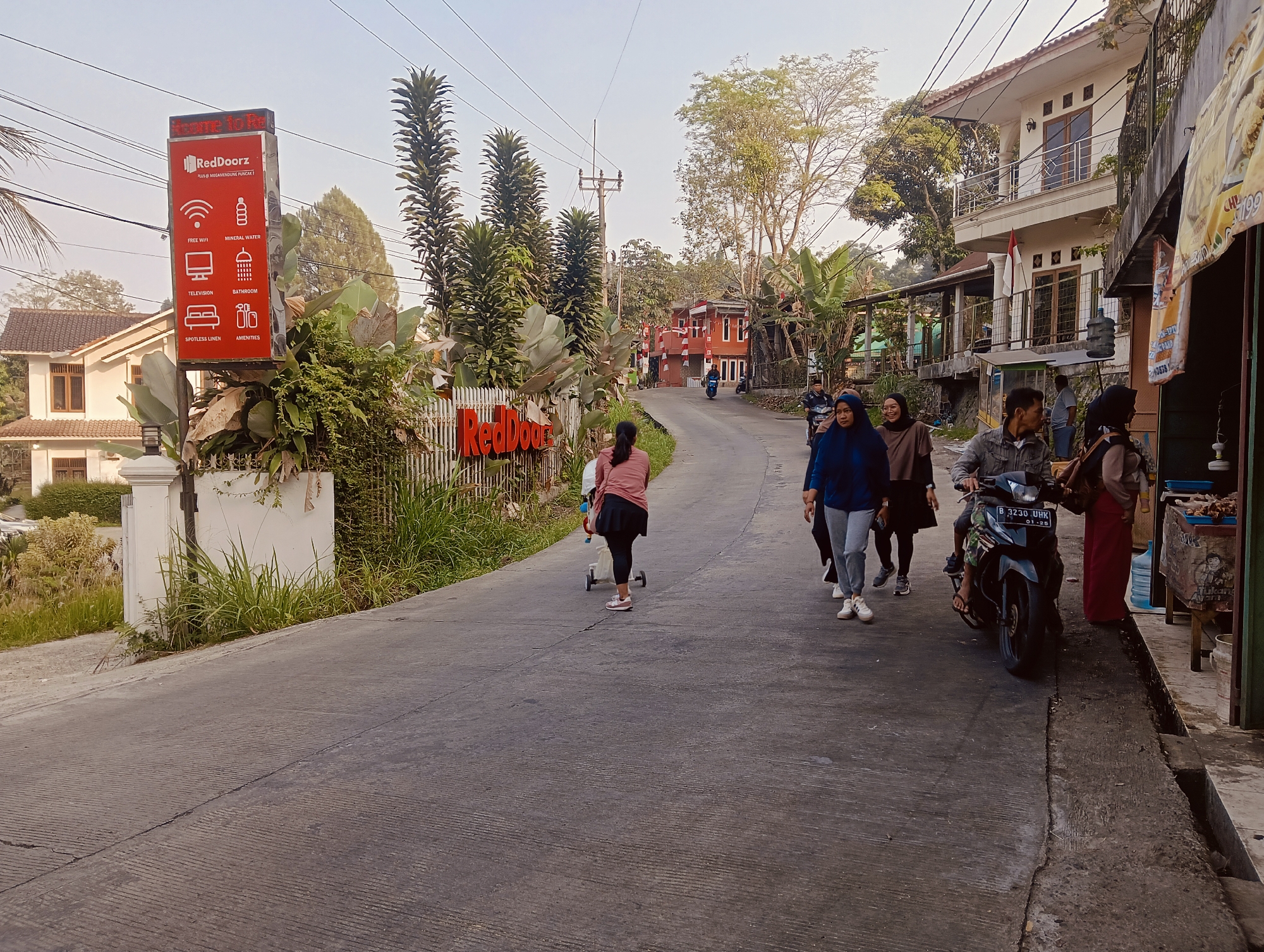 The image depicts a street scene in Megamendung, Indonesia, characterized by a paved road winding uphill. To the left, a prominent RedDoorZ sign stands tall, detailing amenities such as free WiFi, television, mineral water, bathroom, spotless linen, and general amenities. Adjacent to the sign, a white fence and lush greenery flank the road. Further up the street on the left, a modern, multi-story white house with balconies is visible. On the right side of the road, a commercial establishment with a tiled roof and an open storefront is present, featuring goods displayed outside.
Several individuals are captured in the scene. A young girl on a scooter is moving up the road. A group of four women is walking in the same direction, engaged in conversation. A man on a motorcycle is approaching from the background, with another person seated behind him. A woman in a red garment stands near the storefront on the right. The environment suggests a semi-rural or suburban setting with abundant trees and vegetation. The sky is clear, indicating daytime. The road surface shows signs of wear and tear, with visible cracks and parallel grooves.