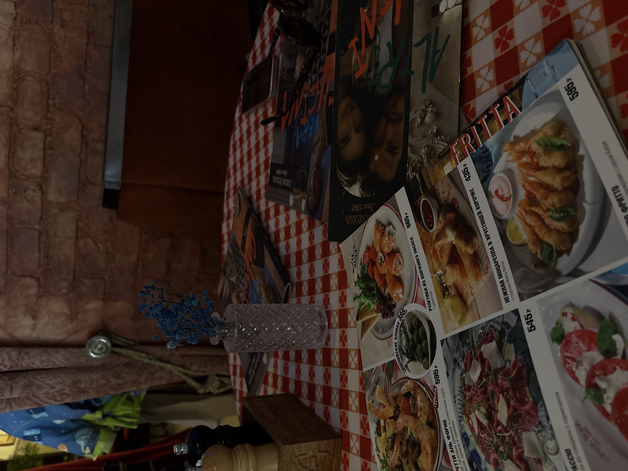The image shows a table with a red and white checkered tablecloth. Several menus are visible, displaying dishes like "Fritto", "Retti Al Forno", and others. These menus, combined with the mention of "Roma since 1998" suggest an Italian restaurant, potentially in Saint Petersburg, Russia (though the image alone cannot confirm location). Other items on the table include sunglasses, magazines, a small vase of blue flowers, and what appears to be a pepper grinder.