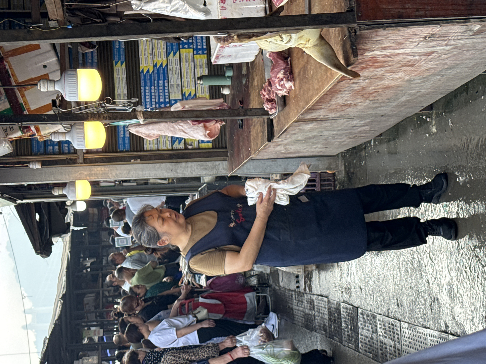A woman, likely a vendor, with grey hair pulled back, stands at a market stall in Jiangjin, China. She is wearing a dark blue apron over a tan shirt and black pants. In her hands, she holds a white cloth. In front of her is a wooden counter laden with cuts of raw meat, including what appears to be a whole pig's head and leg. Behind her, the market extends with a crowded pathway lined with stalls and shoppers. The ground is wet and reflective, suggesting recent rain or cleaning. Overhead, large, bright LED lights illuminate the scene.