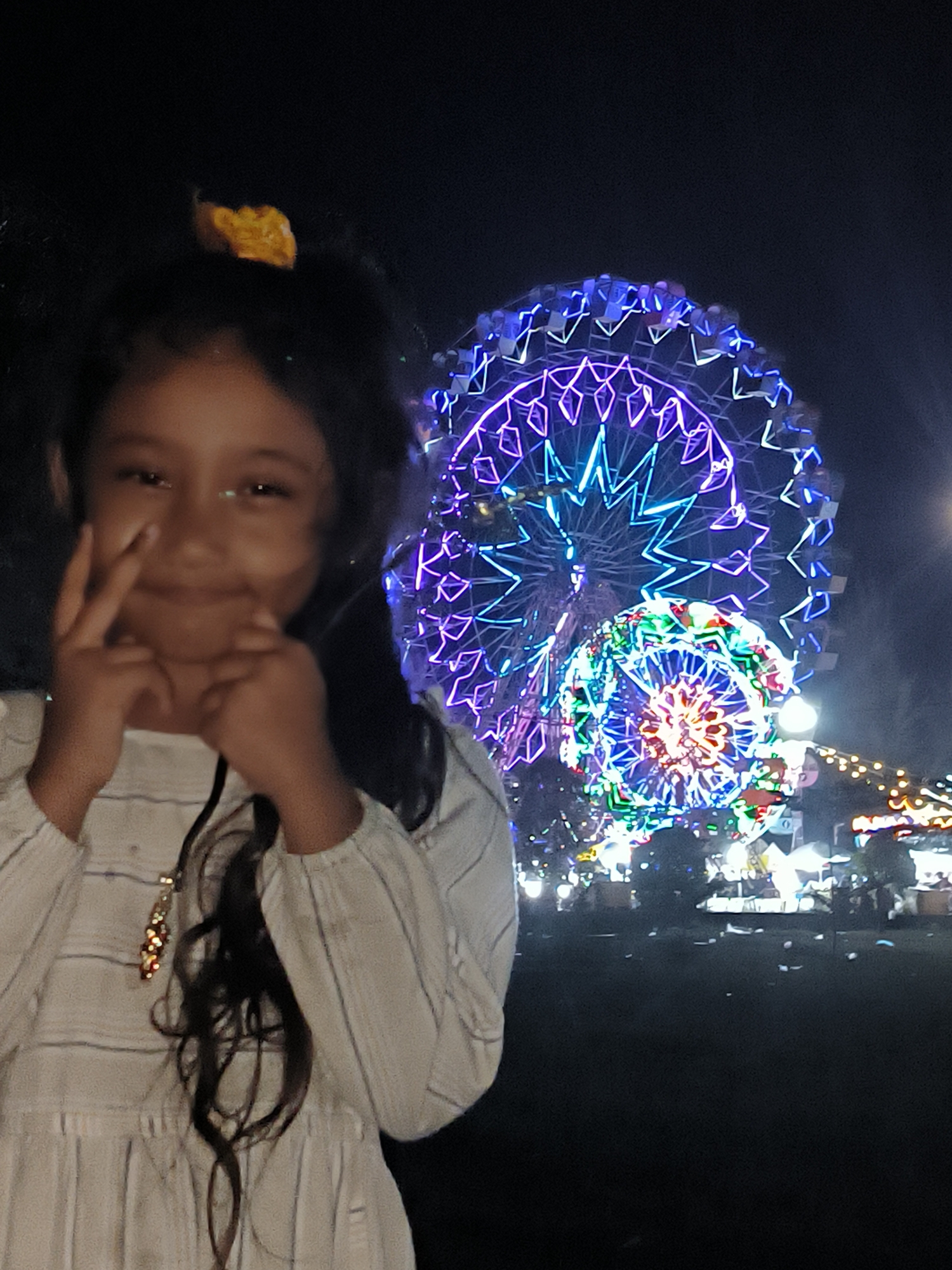 A young girl poses at night in front of a brightly lit carnival. The carnival features two Ferris wheels adorned with vibrant lights in purple, blue, green, and red. In the background, smaller lights indicate other carnival attractions. The girl is dressed in a striped white dress with long sleeves. She has dark hair and is smiling, holding her hands near her face. The scene appears to be in a public outdoor space in Titibaro, Indonesia.
