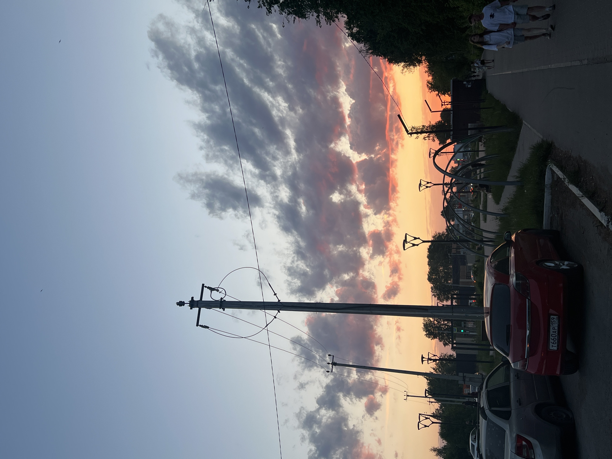 The image shows a street scene in Chaikovsky, Russia at sunset. The sky displays a mix of grey and orange clouds. The street is lined with parked cars, including a red car with a license plate reading "T660AP 159." Overhead power lines are visible. In the background, a walking path with two people is visible. A playground structure is located to the right of the frame. A building with a green light showing through the trees.
