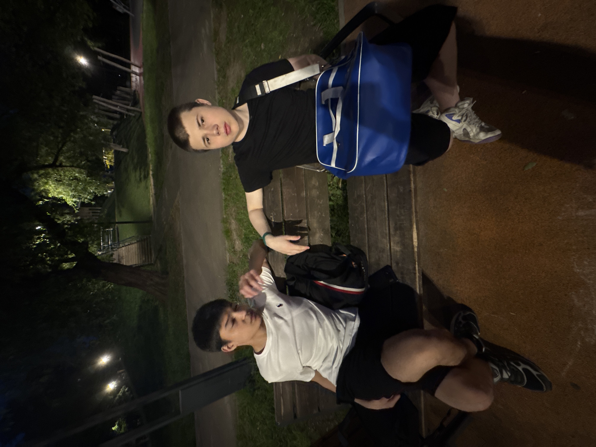 Two young men are sitting on a park bench at night. The first man is wearing a black shirt and black shorts, and white and blue sneakers. He has a blue duffel bag on the bench next to him. The second man is wearing a white shirt and black shorts and black and white sneakers. A black backpack is sitting next to him on the bench. The background shows a dimly lit park with trees and a wooden structure in the distance. Based on the location and information, the scene is likely in a park in Moscow, Russia.