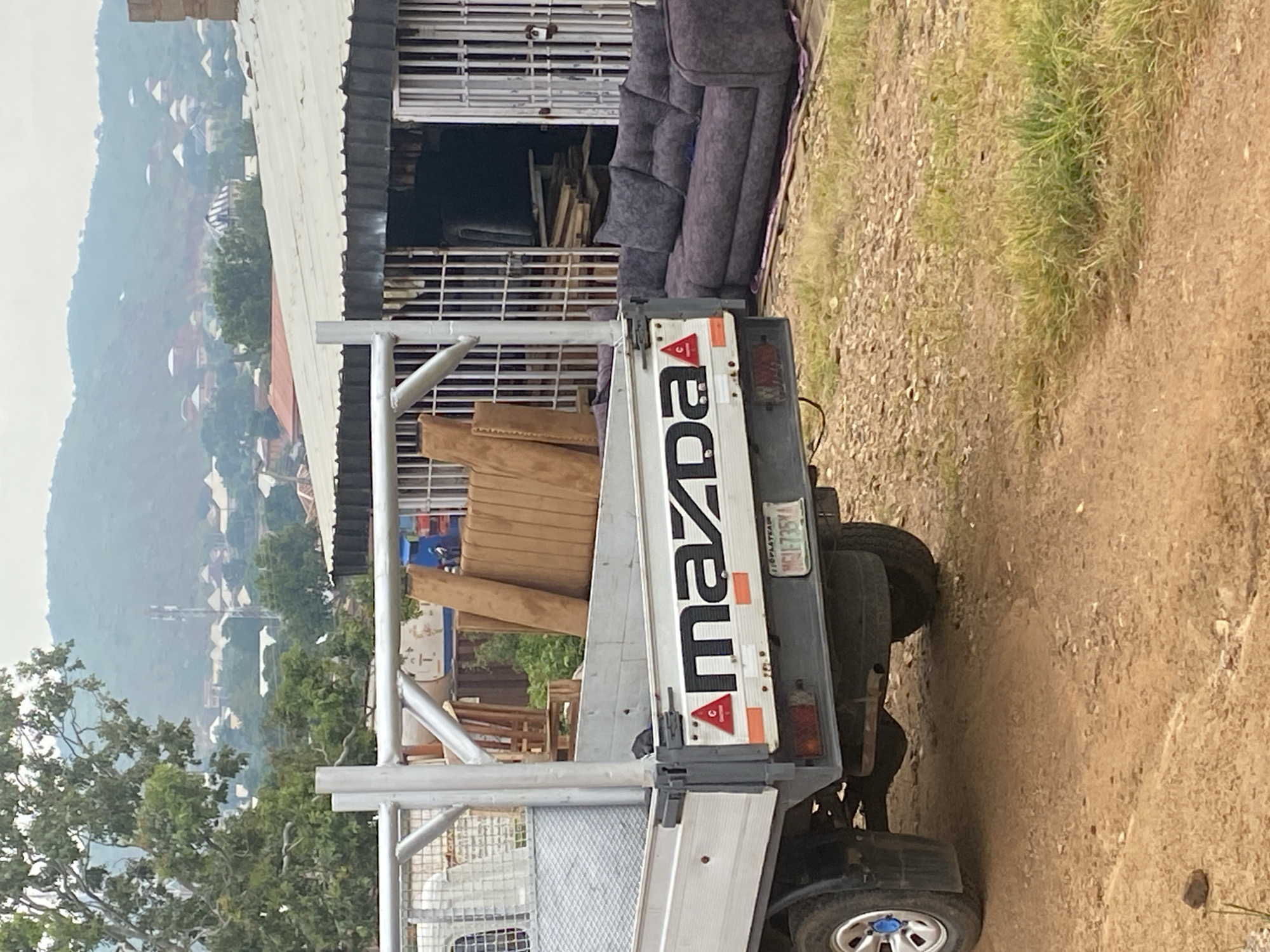 In Jos, Nigeria, a flatbed truck with the "Mazda" logo on its tailgate is parked on a dirt road. The truck bed contains several upholstered furniture items, including what appears to be parts of a couch or chair. Adjacent to the truck, on the right side of the image, is a gray, tufted sectional sofa. Behind the truck and sofa is a building with a corrugated metal roof and barred windows, suggesting it may be a shop or workshop. In the background, a hazy view of a residential area with houses and vegetation is visible on a hillside under a cloudy sky.