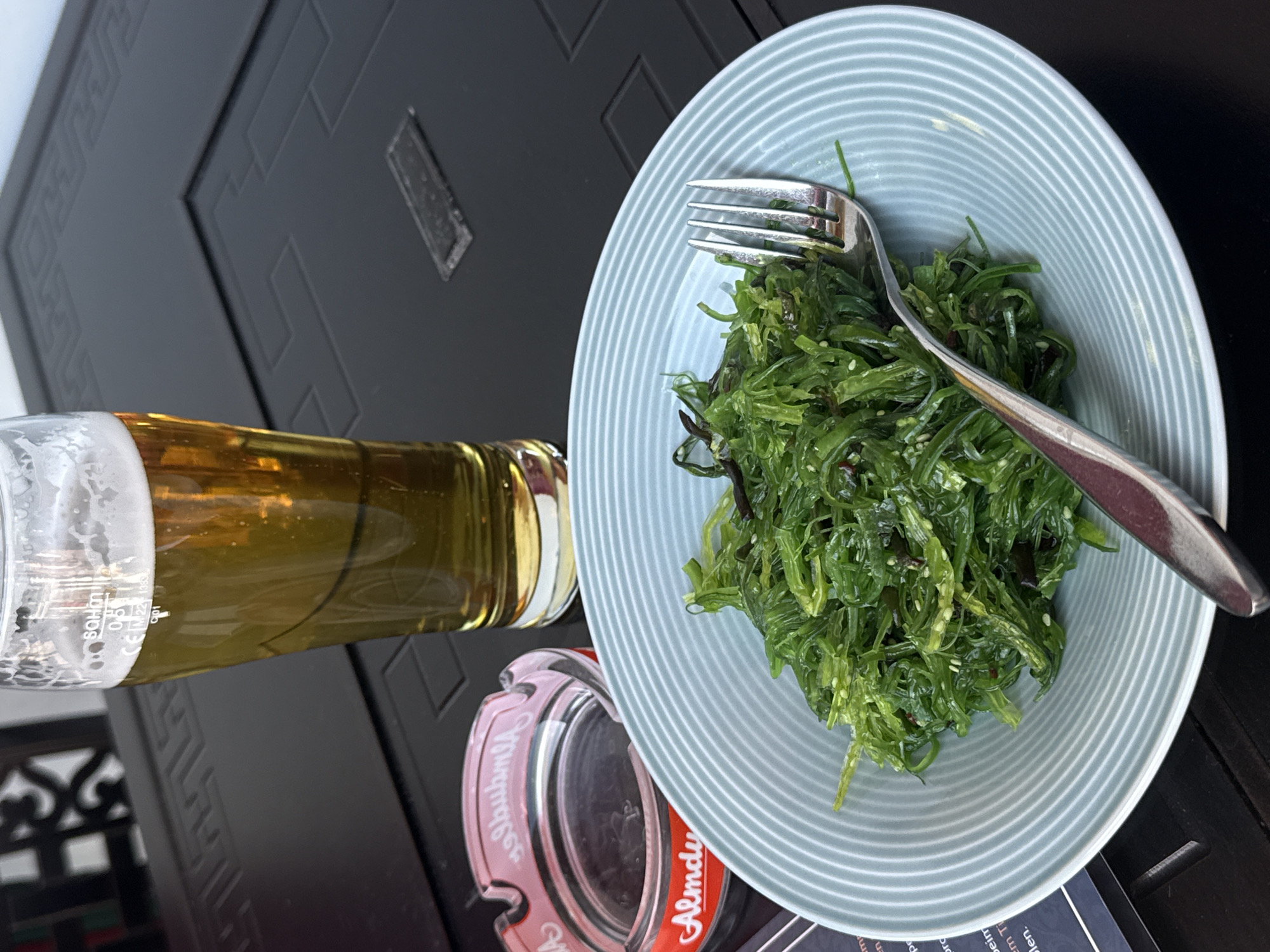 The image shows a plate of seaweed salad with a fork on a dark table. A bottle of beer and an ashtray are next to the plate. The location is Hainburg an der Donau, Austria.