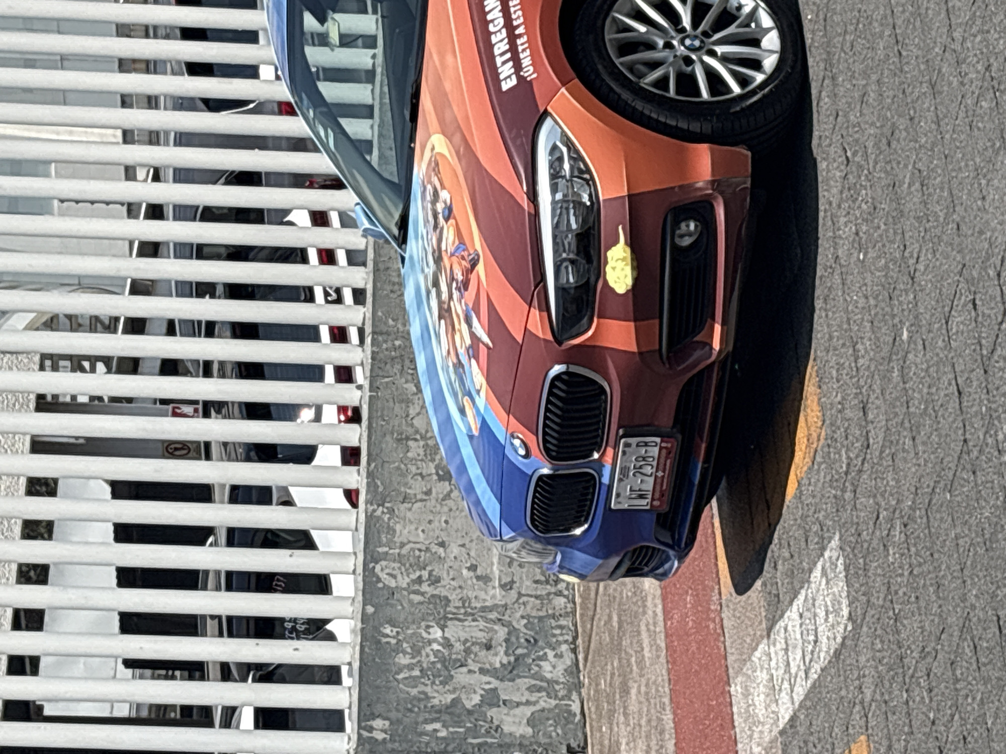 The image depicts a BMW Z4 with a custom paint job, mostly in shades of blue and orange, with the license plate "LWF-258-B". The car is parked near a white metal fence, with the vehicle's front end angled towards the camera. The ground surface is paved, with red and white painted traffic markings. The picture was taken in the city of Huixquilucan de Degollado, Mexico.