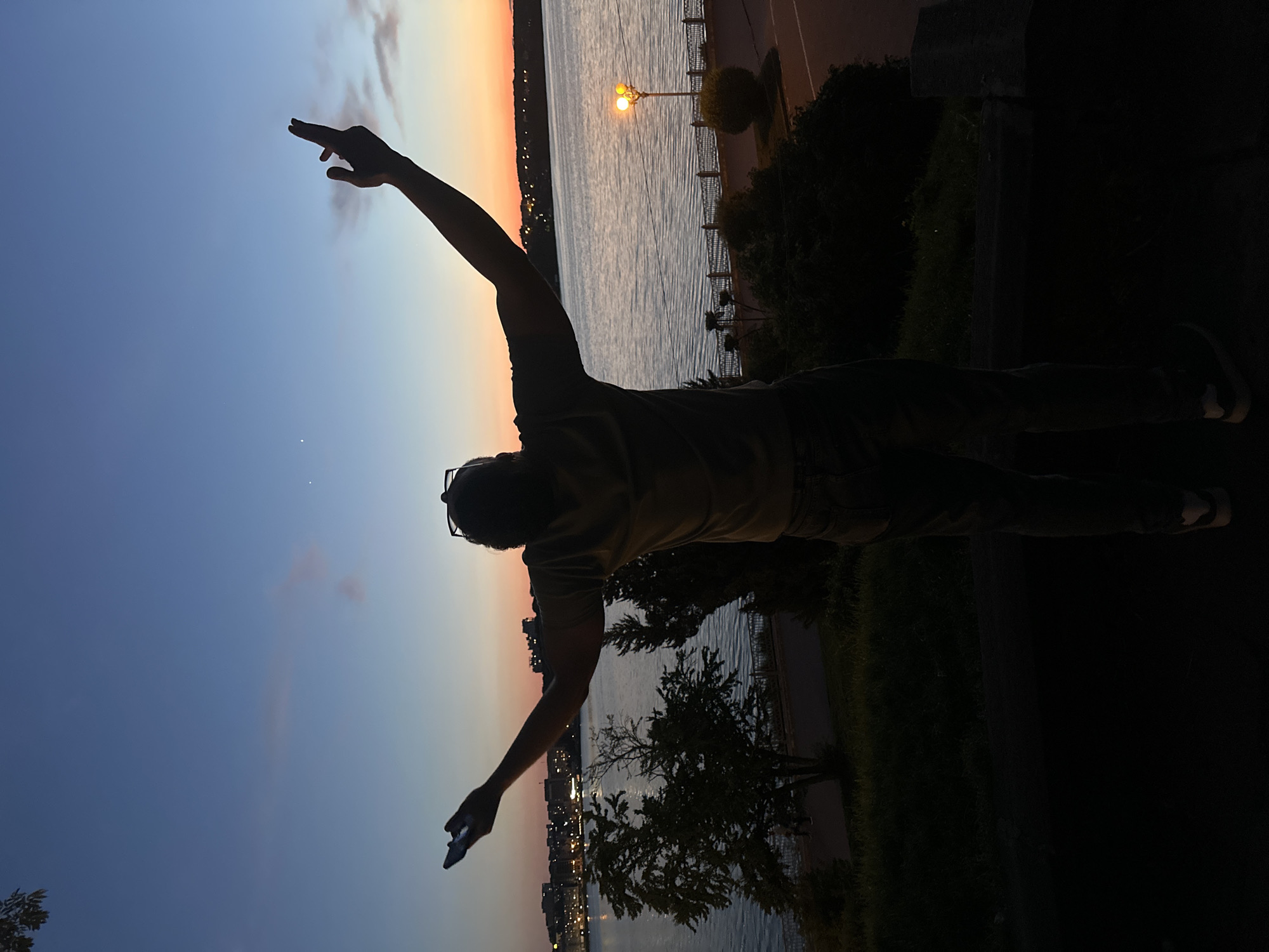 The image depicts a solitary individual captured in silhouette against a sunset sky, overlooking the Ternopil Pond in Ternopil, Ukraine. The scene is set at dusk, with the sky transitioning from deep blue at the zenith to pale blue, orange, and pink hues at the horizon. Two faint white celestial bodies are visible in the upper left sky.
The central subject, a person, stands with their back to the viewer, arms outstretched upwards and outwards. They appear to be wearing a t-shirt, jeans, and sneakers, with sunglasses perched on their head. A smartphone is held in their left hand, angled upwards.
In the mid-ground, the Ternopil Pond extends across the frame, reflecting the twilight sky with ripples visible on its surface. Across the pond on the left, a cityscape with illuminated buildings is visible on the distant bank. To the right, the far bank is tree-lined and also shows faint lights along its horizon.
Along the near side of the pond, below the level of the standing person, there is a paved promenade or embankment bordered by a dark metal railing. Several dark-silhouetted trees and shrubs are interspersed along this area. A single streetlamp with a bright, warm yellow-orange glow illuminates a section of the path on the right side of the frame. The foreground where the person stands is dark and shadowed, consisting of ground cover and a visible concrete ledge or barrier. The elevated vantage point overlooks the promenade and the pond.