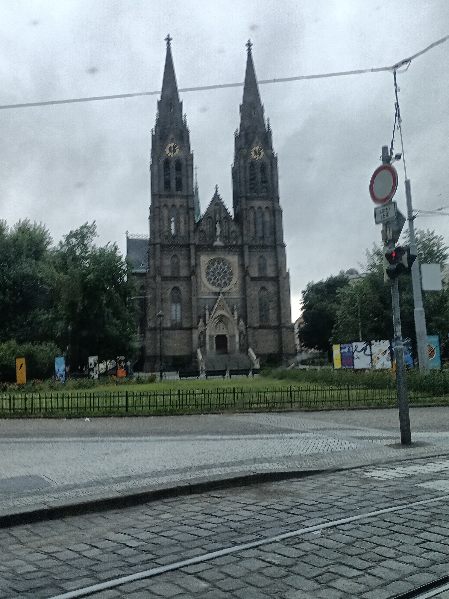The image captures a street view in Prague, Czechia, focusing on the prominent gothic spires of the Basilica of St. Peter and St. Paul at Vyšehrad. The sky is overcast, contributing to a subdued lighting condition. In the foreground, a cobblestone street with tram tracks is visible. A black metal fence separates the street from a grassy area, beyond which the impressive stone facade of the basilica stands tall with its twin spires. The basilica features arched windows and a large rose window on its front. To the right of the basilica, a traffic light is visible, showing a red signal. Behind the fence, several trees and some posters are also present. The overall scene is a static, architectural-heavy cityscape.