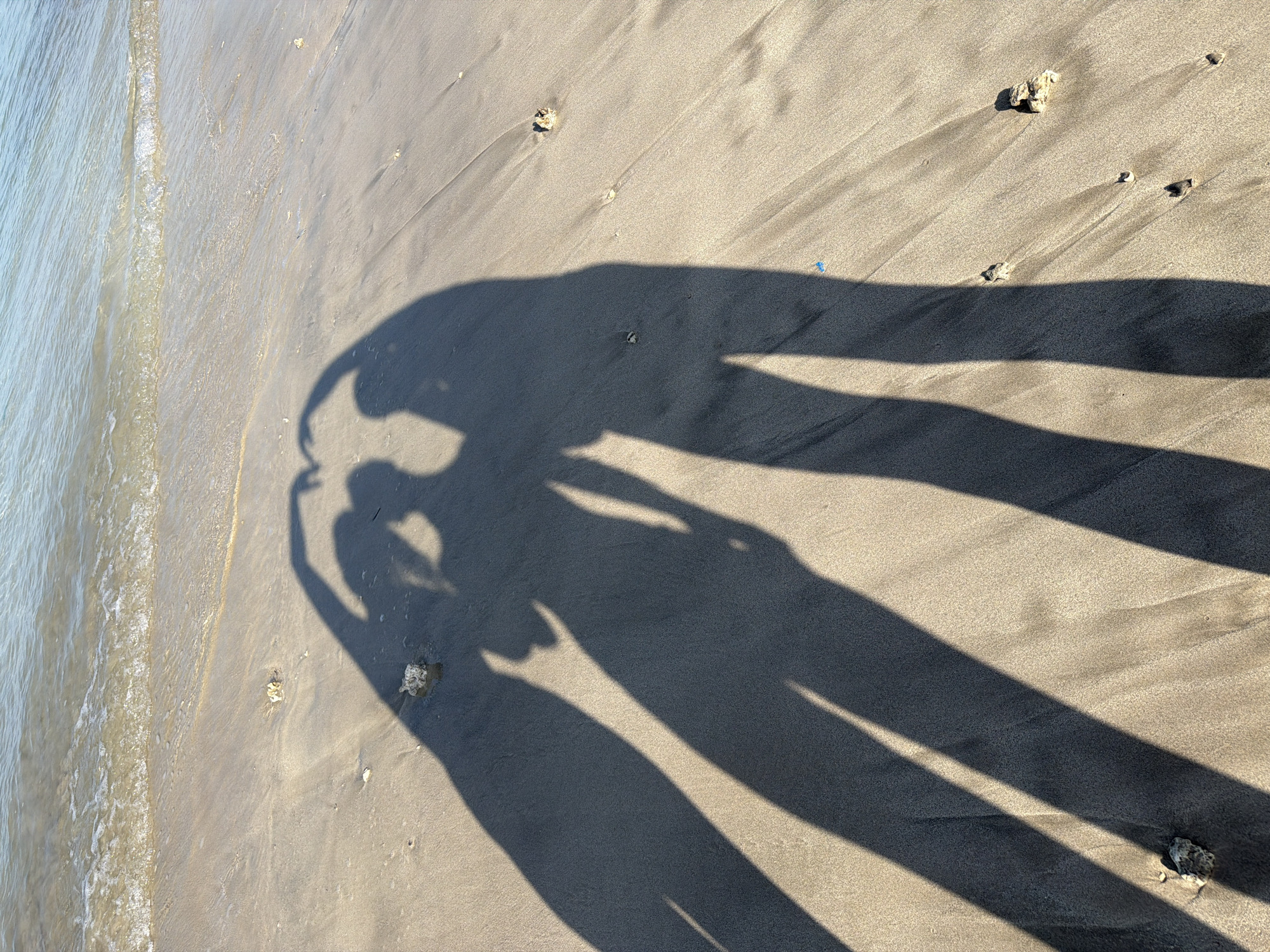 The image depicts a beach scene in Kuta, Indonesia. The sandy shore is visible, with water lapping at the edge of the sand on the left side of the image. Shadows of two people taking a selfie are cast on the sand. Small pebbles and pieces of debris are scattered across the beach.