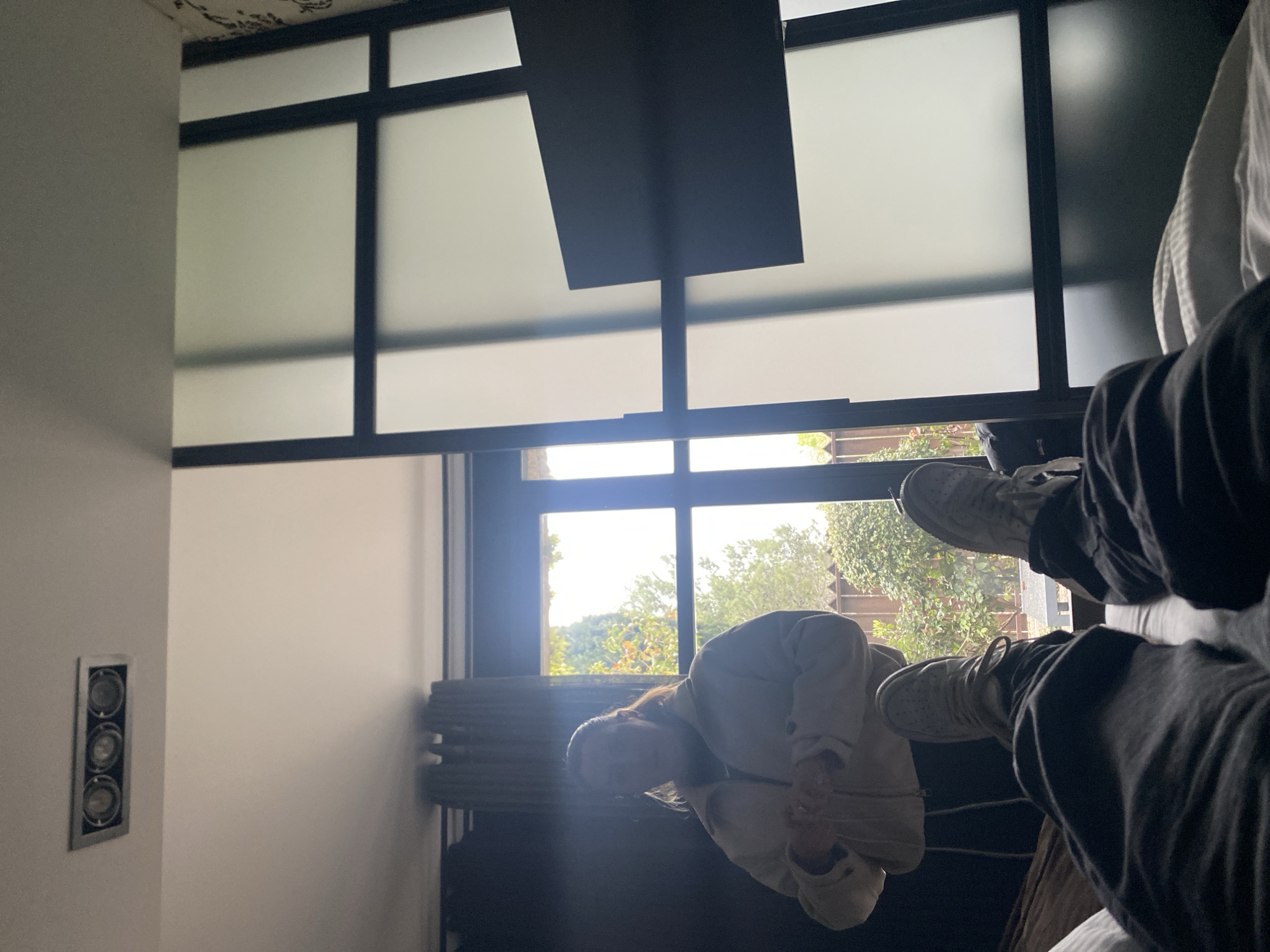 A person is indoors, sitting in a chair with their legs up, visible against a window. The window provides a view of trees and possibly a landscape outside. Another person is visible in the lower half of the image, possibly crouching near the chair. The interior space features a window with a grid-like pattern, a ceiling light, and a wall with a light control panel. The image does not provide enough information to confirm if the location is in Carcassonne, France.