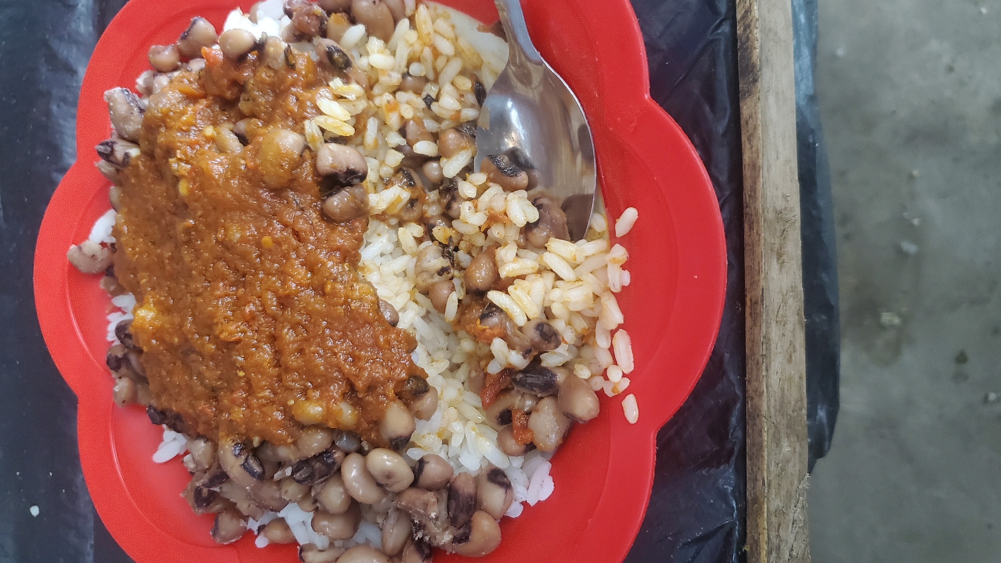 A top-down shot of a red, heart-shaped plastic plate filled with black-eyed peas and white rice. A thick, reddish-brown stew is spooned over a portion of the rice and peas. A silver spoon rests on the right side of the plate, partially submerged in the rice. The plate sits on a black surface, possibly a plastic bag, which is in turn resting on a wooden surface. The background is blurred and appears to be a concrete or dirt surface. The overall lighting suggests an outdoor or dimly lit indoor setting. The scene is a close-up of a meal, with no people or clear geographical markers visible to indicate a specific location in Jalingo, Nigeria.